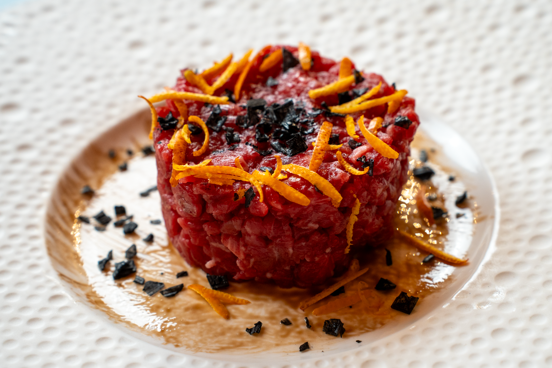 Tartare in un stampo e decorata con scorze di agrumi e pepe, su un piatto bianco con decorazioni scentifiche.