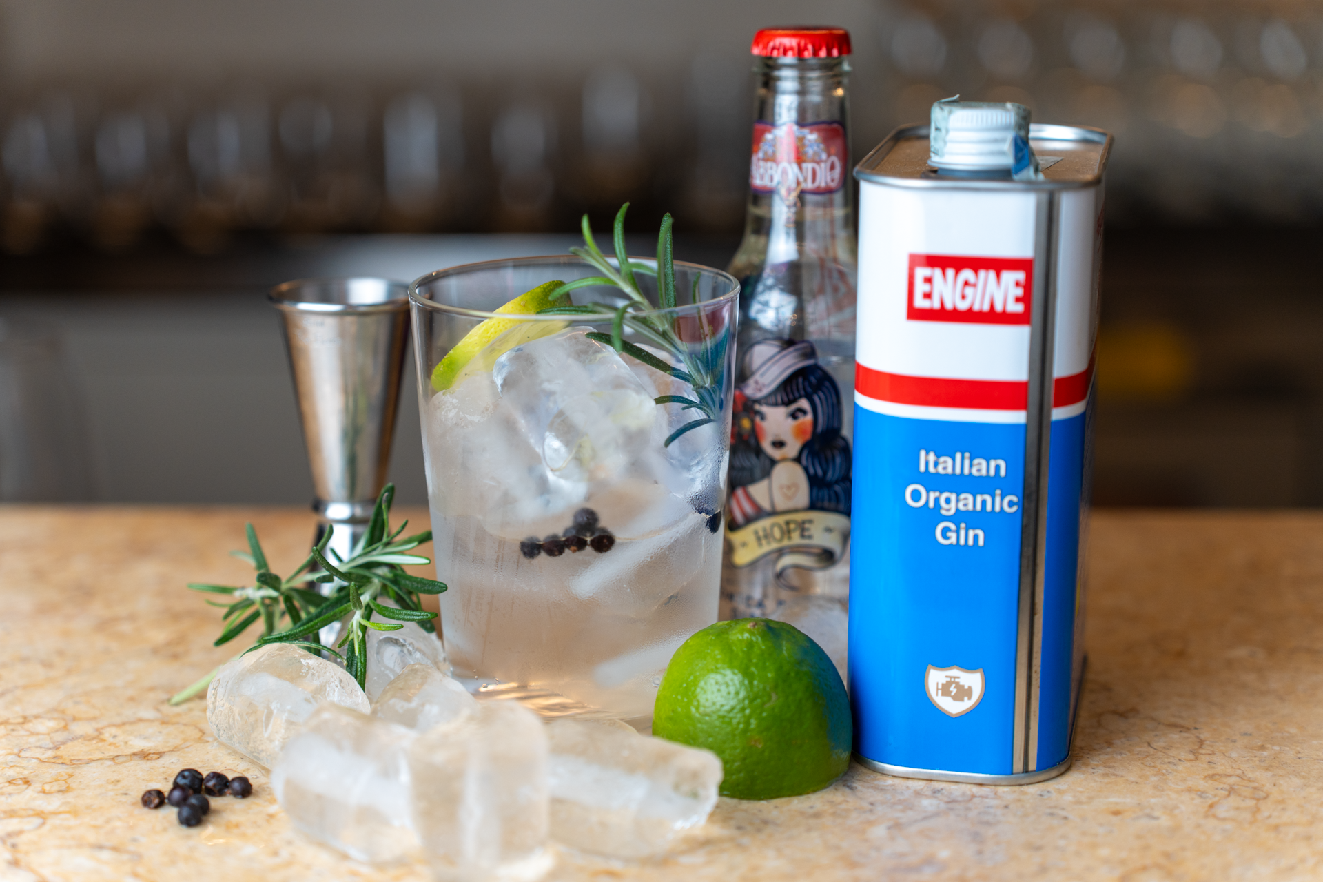Cocktail con ghiaccio, limone e rosmarino, con una bottiglia di gin biologico italiano, una scatola di combustibile per fuochi e alcuni lime, decorato con rosmarino e pepe di Sichuan.