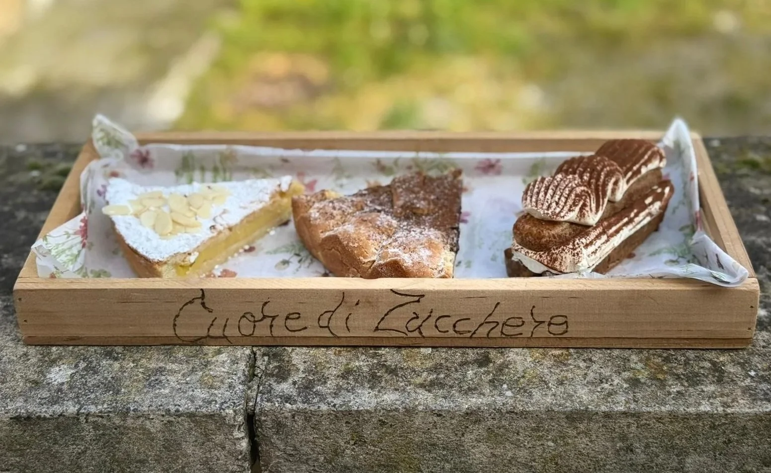 Tre dolci su un vassoio di legno con scritto Cuore di Zucchero, posato su una superficie di pietra. Uno è una fetta di torta al limone con mandorle, un altro è una fetta di torta alla frutta, e l'ultimo è una torta al cioccolato