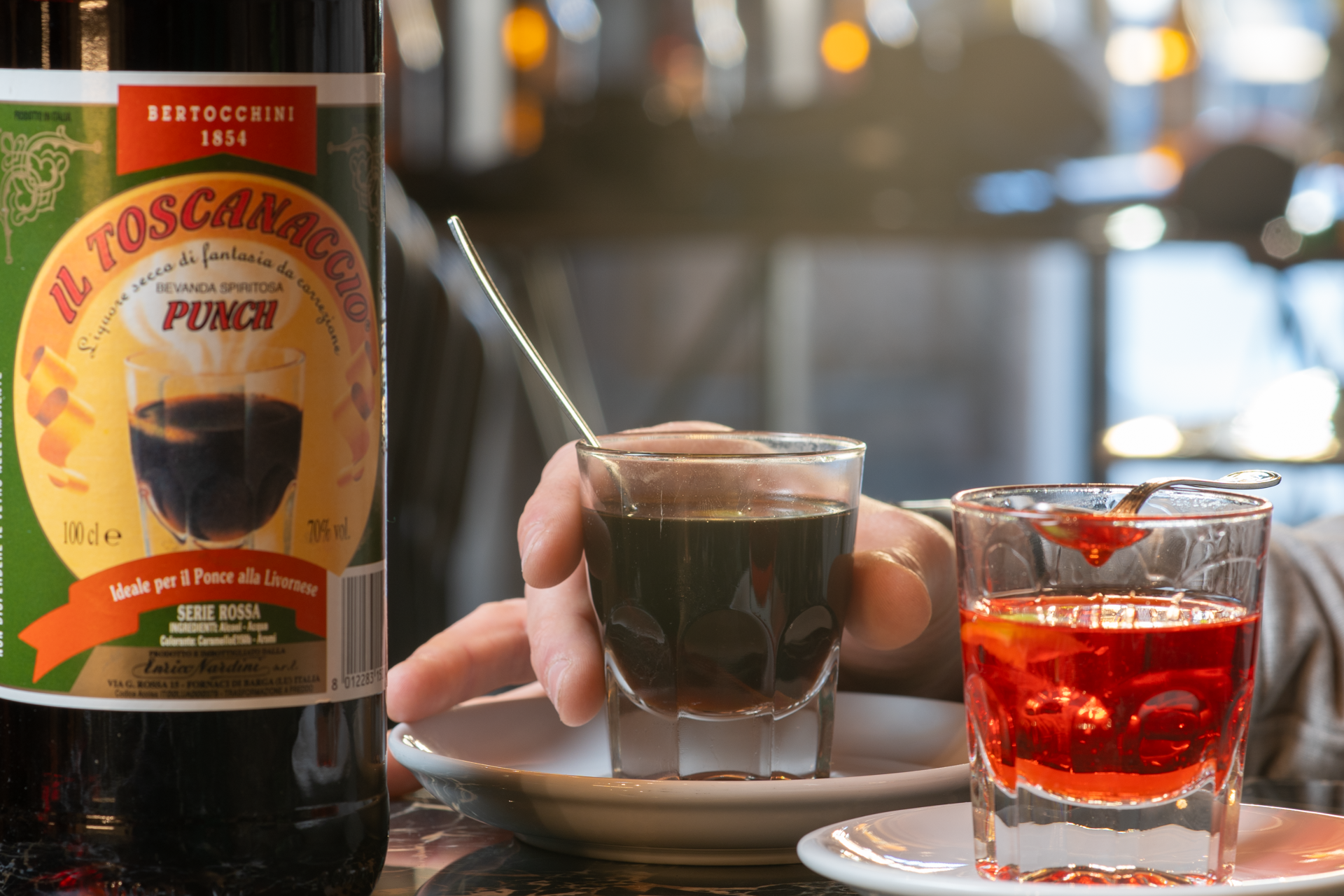 Primo piano di tre bicchieri con liquori diversi, uno nero, uno rosso con sciroppo, e uno trasparente, con una mano che li tiene, su un piatto bianco, con una bottiglia di drink sullo sfondo.