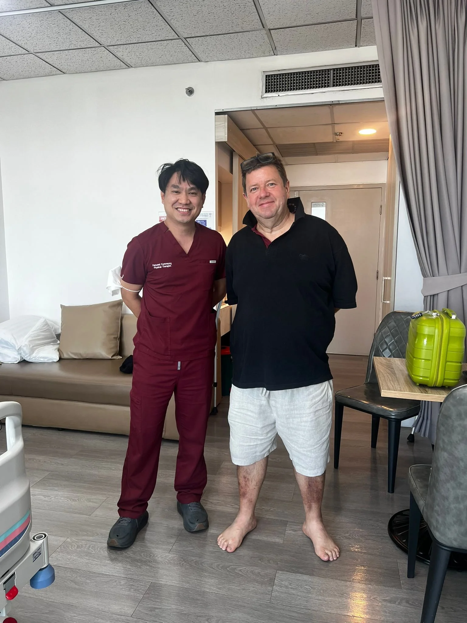 A man in casual clothing with glasses on his head stands next to a healthcare worker in medical scrubs, both smiling inside a medical facility, with some medical equipment and furniture around.