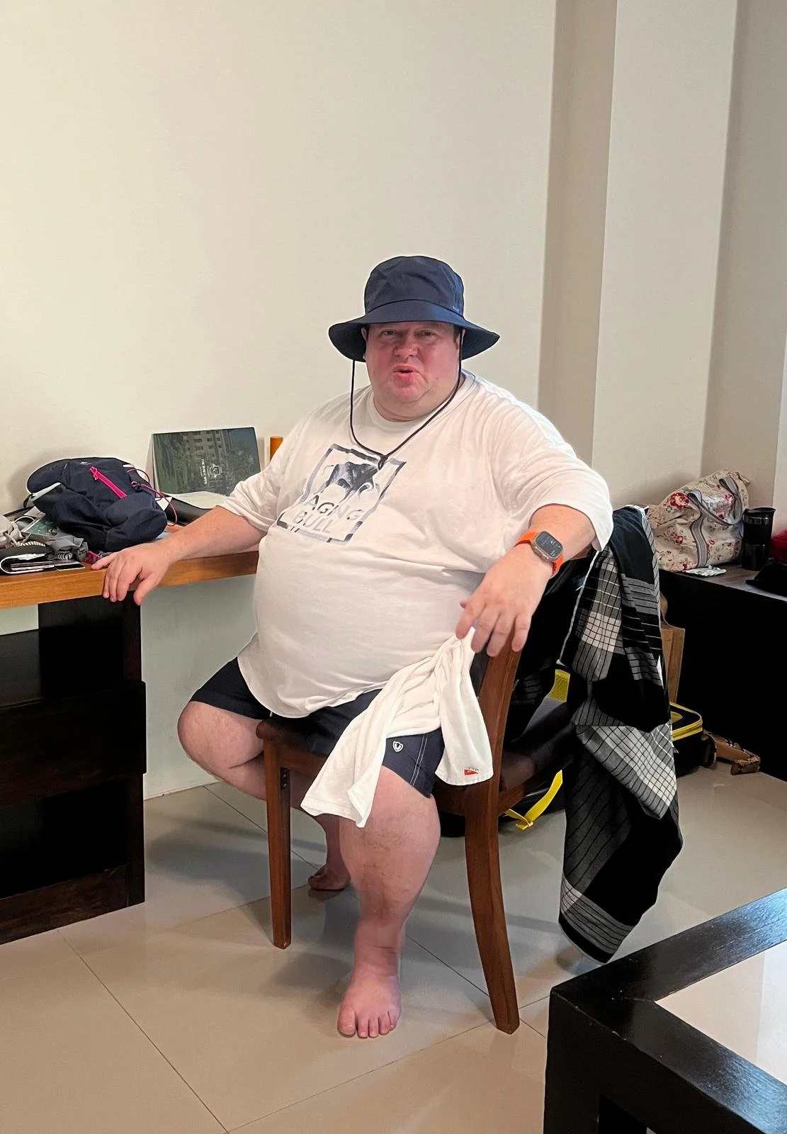 A man sitting on a wooden chair in a room, wearing a wide-brimmed hat, white T-shirt, shorts, and a watch, with a towel draped over his lap, surrounded by personal belongings and a bag in the background.