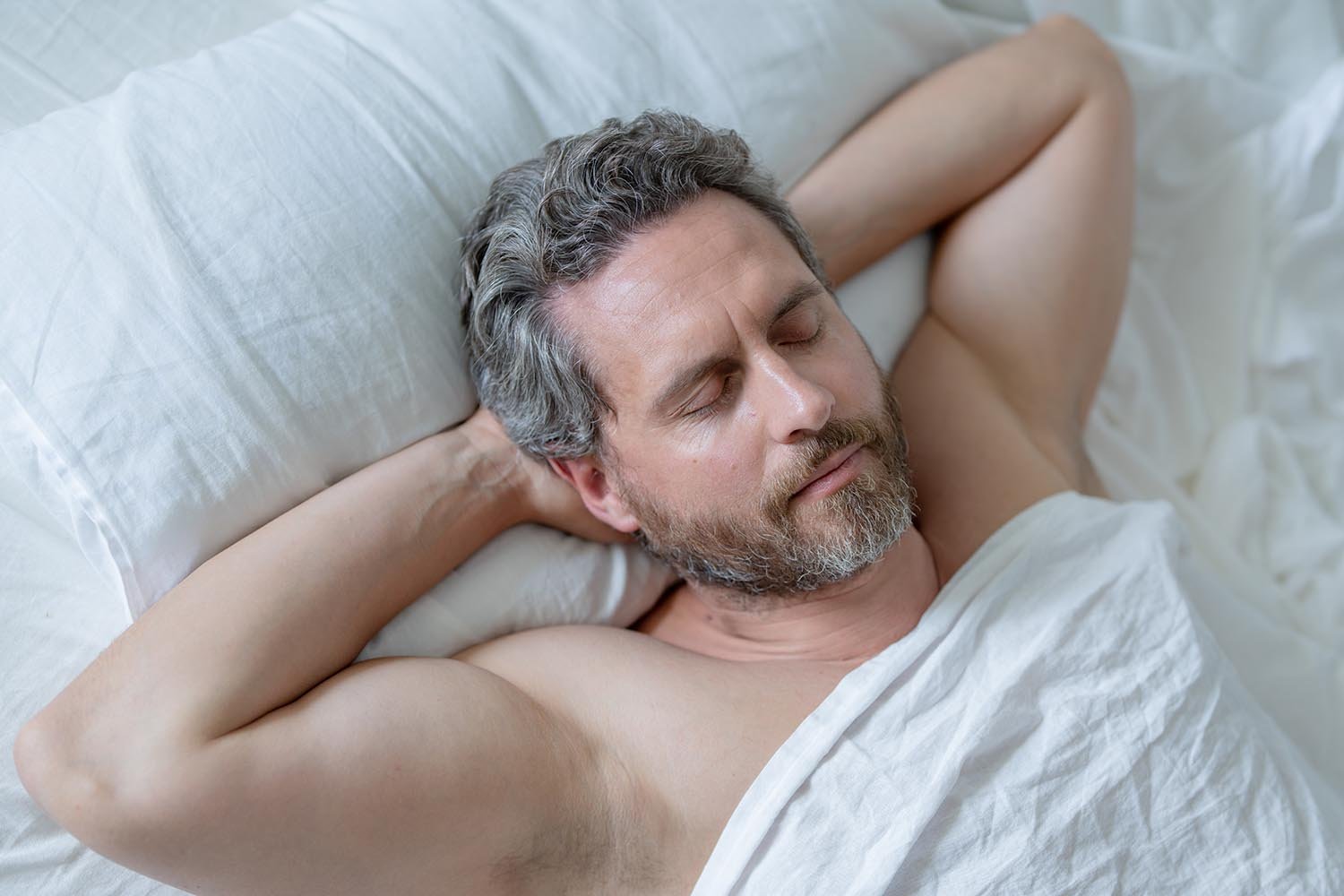 A middle-aged man with gray hair and beard sleeping peacefully in bed with a white pillow and blanket.