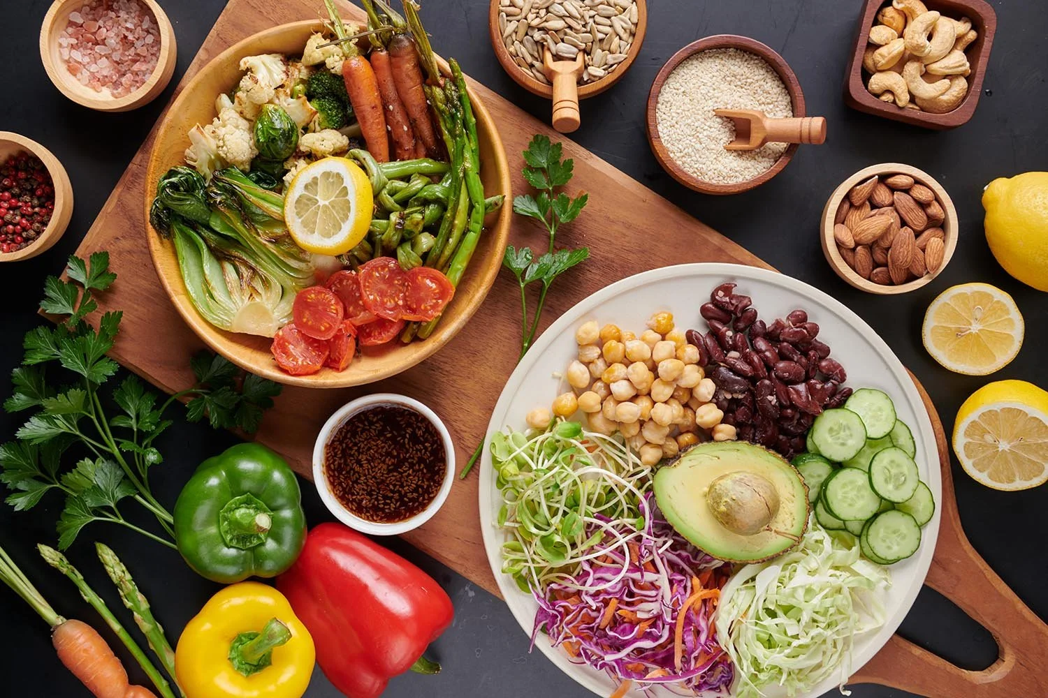 Assorted fresh vegetables, fruits, nuts, seeds, and salad ingredients on a black surface, including tomatoes, cucumbers, avocado, carrots, peppers, cashews, almonds, sunflower seeds, sesame seeds, pink salt, lemon slices, and a salad bowl with mixed vegetables and chickpeas.