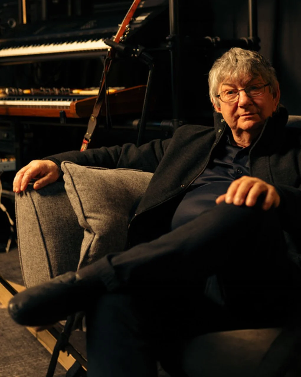 An older man with glasses sitting on a gray sofa, with a grand piano and a guitar in the background.