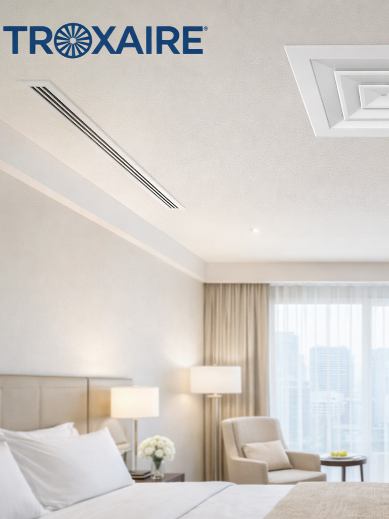 Interior of a hotel room with a bed, lamps, and a sitting area featuring an armchair and a window with curtains, branded with the Troxaire logo in the top left corner.