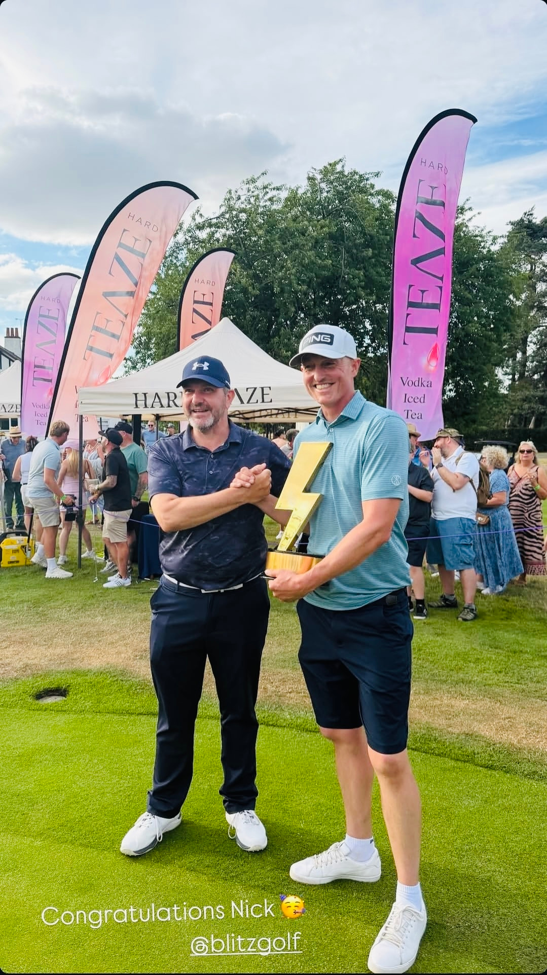 Two men at a golf event, one holding a yellow lightning bolt trophy. Both are smiling, one wearing a navy cap and the other a white cap. Behind them is a crowd, tents, and pink banners with the words 'Teez Hard' and 'Vodka Iced Tea.'
