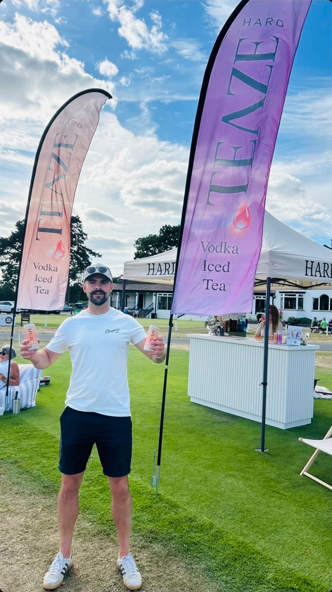 A man standing outdoors on grass, holding two cans of vodka ice tea, with two tall pink banners that say "Hard VEZE Vodka Iced Tea" behind him. There are tents and other people in the background.