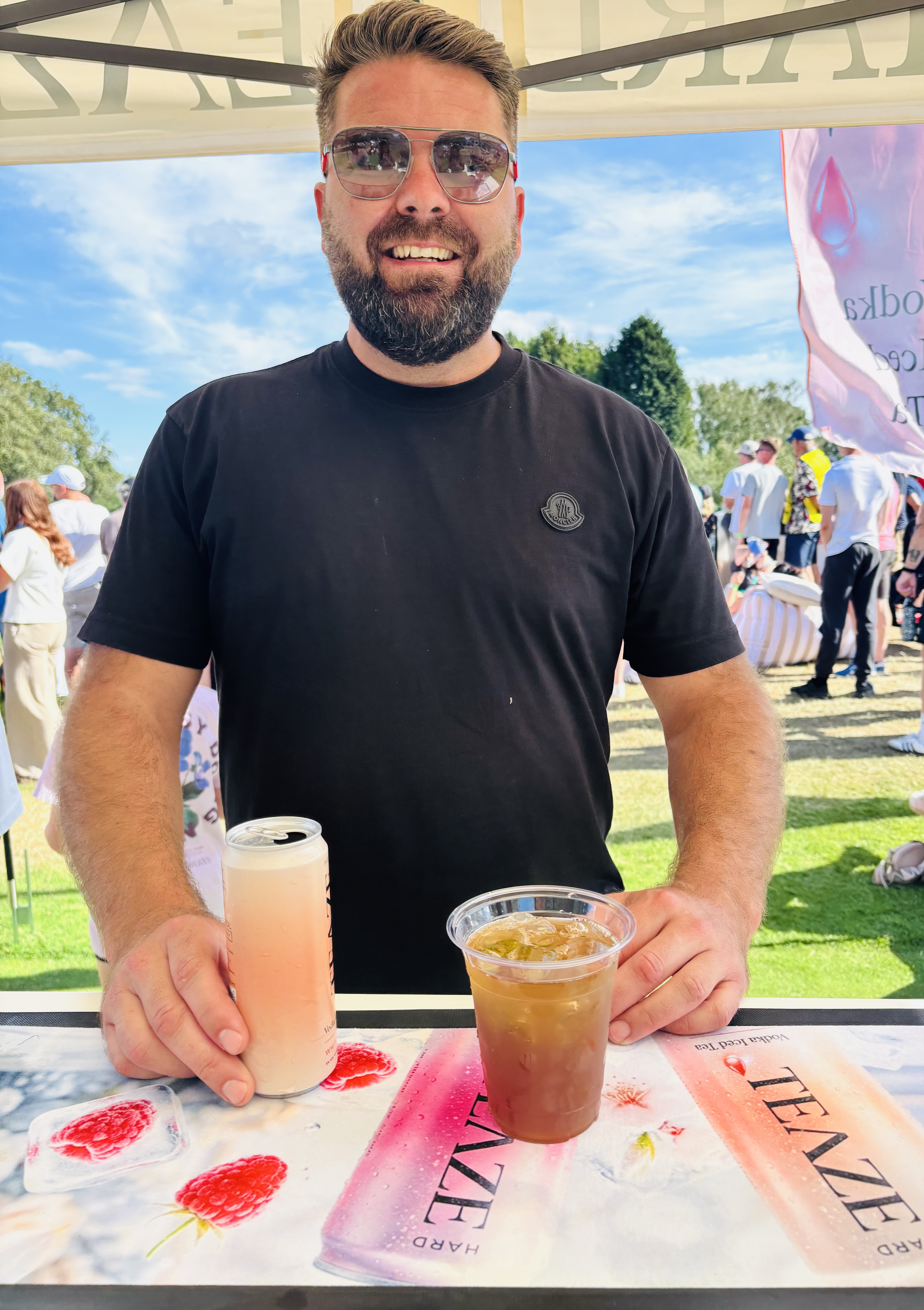 A man with sunglasses, a beard, and a black t-shirt smiling at the camera while holding a Hard Teadrink in each hand, one with a pale pink beverage and the other with a darker beverage, at an outdoor event with a crowd and blue sky in the background.