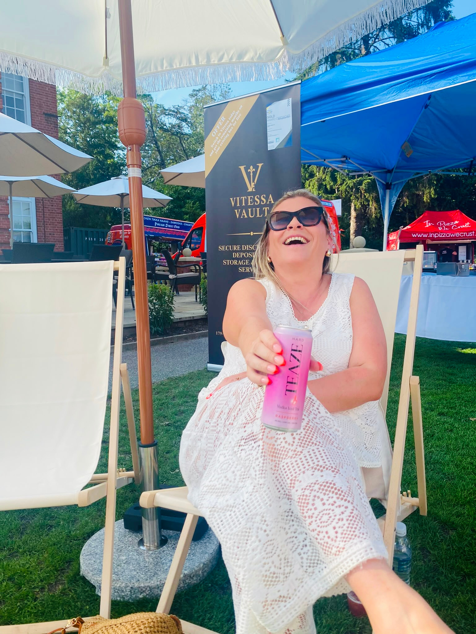 A woman wearing sunglasses and a white dress sitting outdoors at a table with a pink drink, smiling and reaching toward the camera, with umbrellas and a sign for Vitessa Vault in the background.