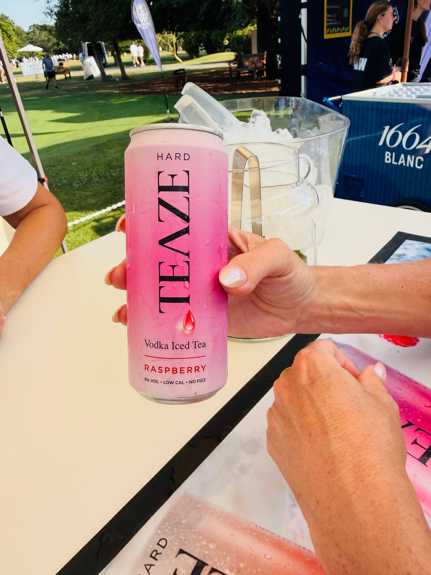 A hand holding a pink can of hard vodka iced tea with raspberry flavor, labeled as 5% alcohol, low calorie, and no fizz. In the background, there are empty glasses, a water cooler, and an outdoor park scene with people walking and trees.