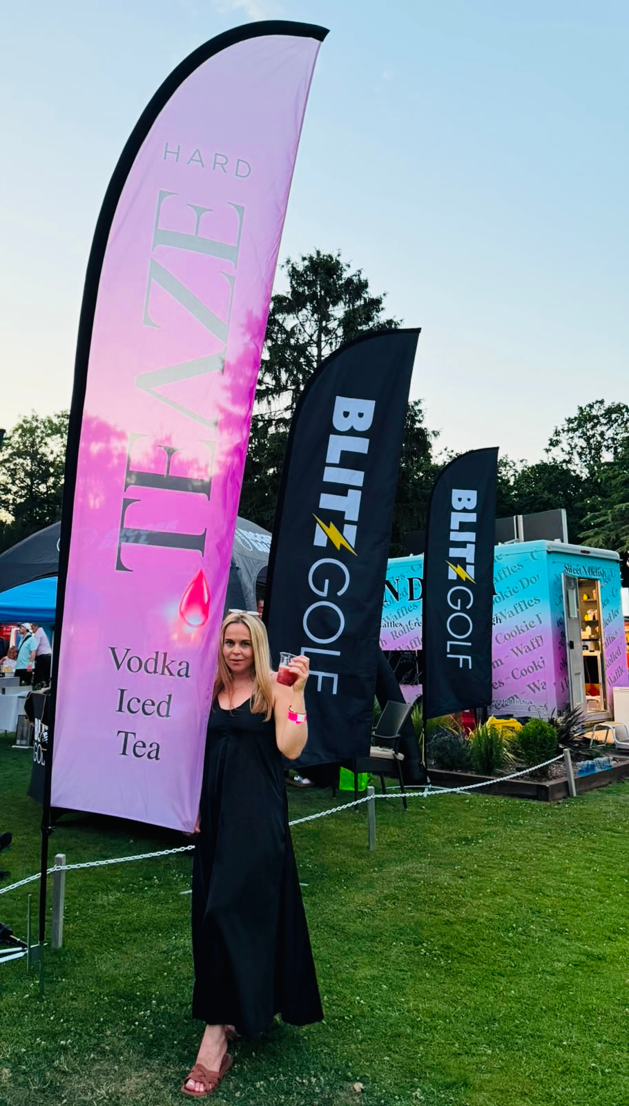 A woman in a black dress holding a drink stands in front of flags advertising vodka iced tea and Blitz Golf at an outdoor event during sunset.