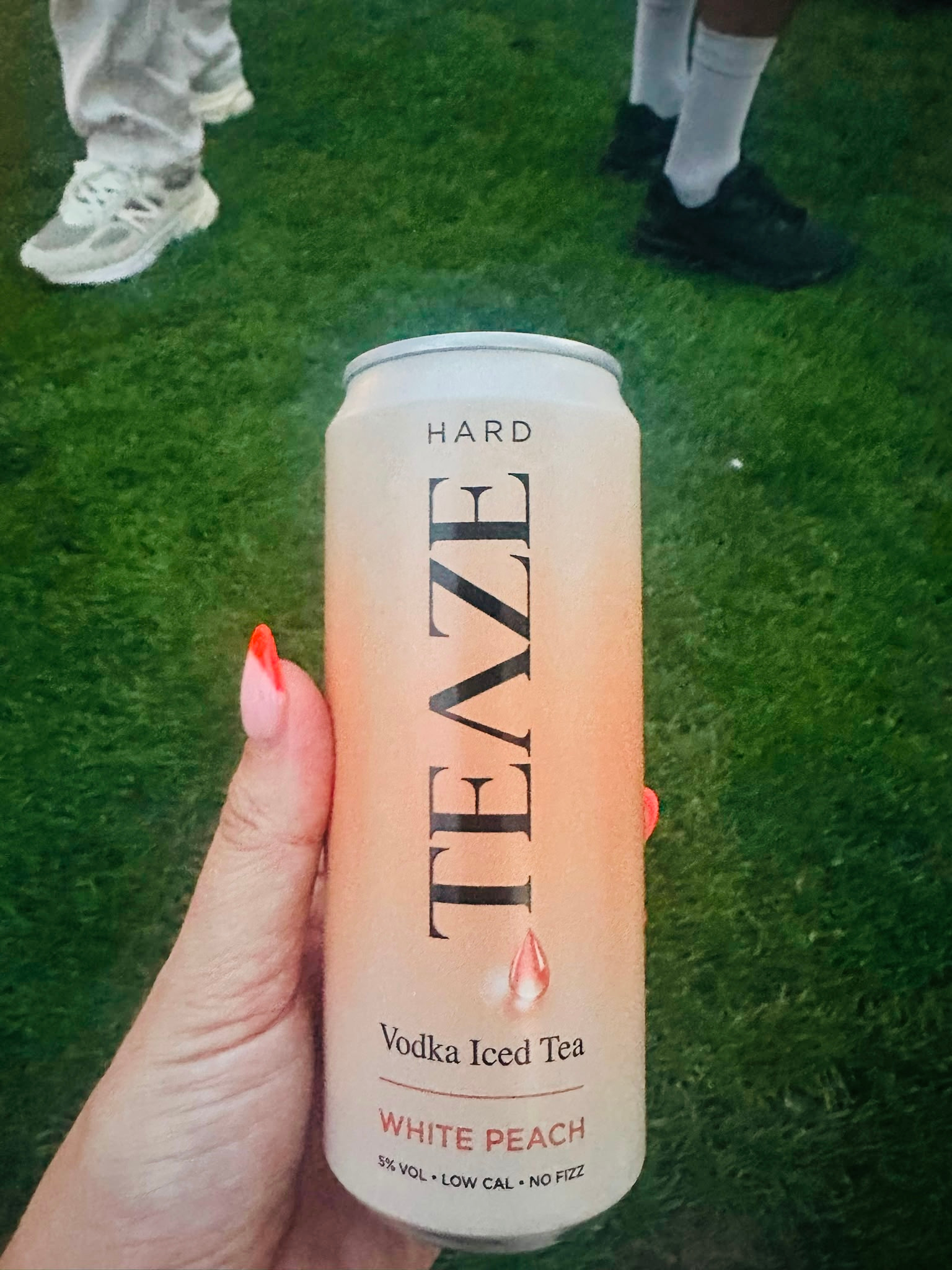 A hand holding a can of White Peach Vodka Iced Tea against a green grass background, with two people standing nearby wearing white and black shoes.