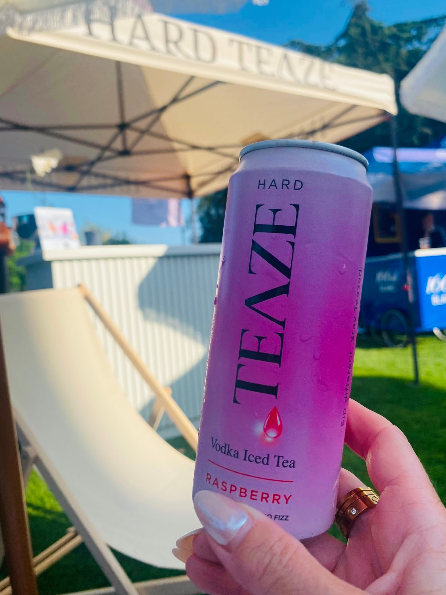 Close-up of a hand holding a pink can of vodka iced tea with raspberry flavor, under an outdoor patio umbrella marked 'GARDEN TEA' on a sunny day.