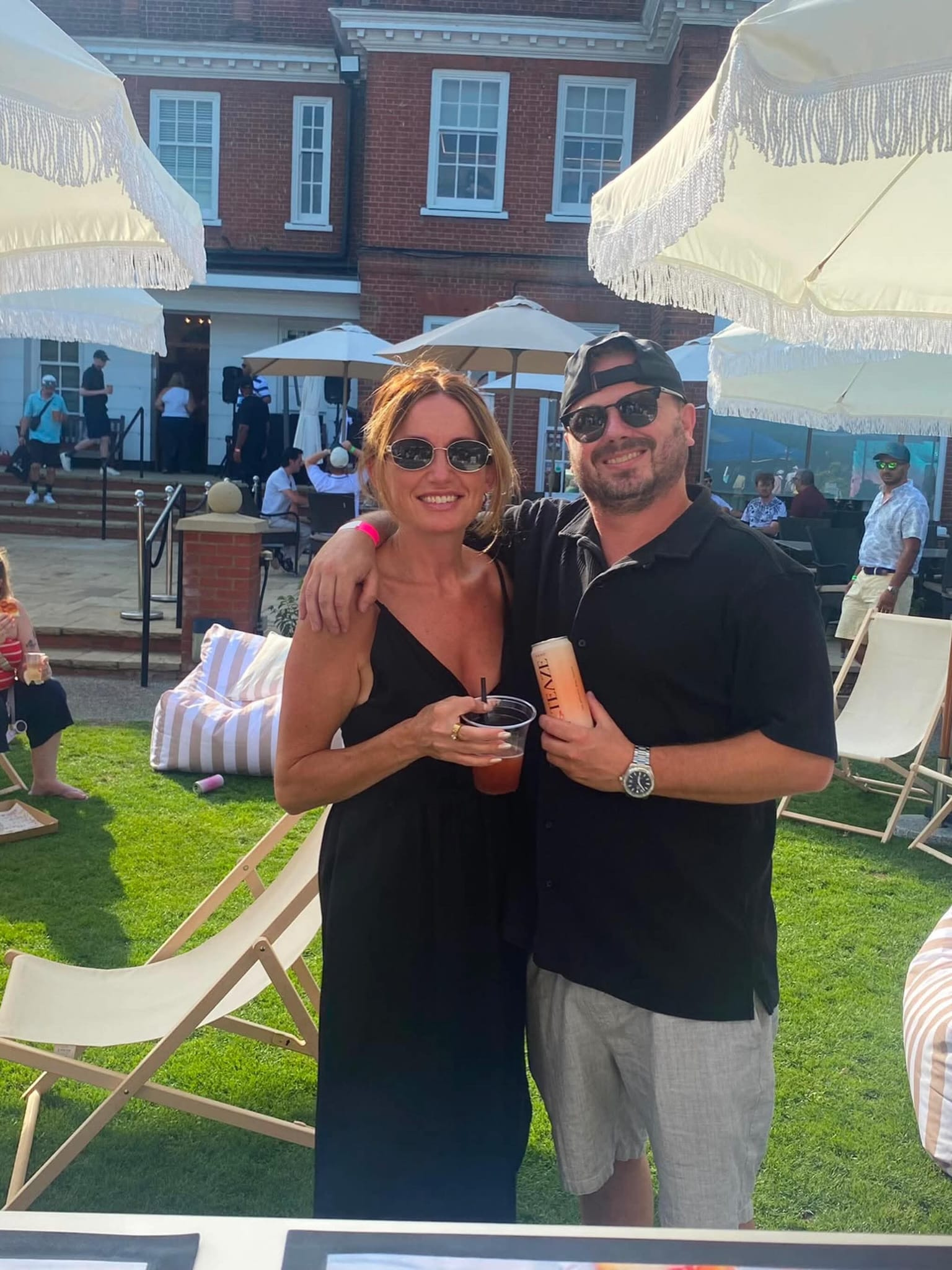 A smiling couple taking a selfie at an outdoor event. The woman is wearing sunglasses and a black dress, holding a drink. The man is wearing a black shirt, a backward cap, and sunglasses, holding a drink. There are umbrellas, lounge chairs, and other