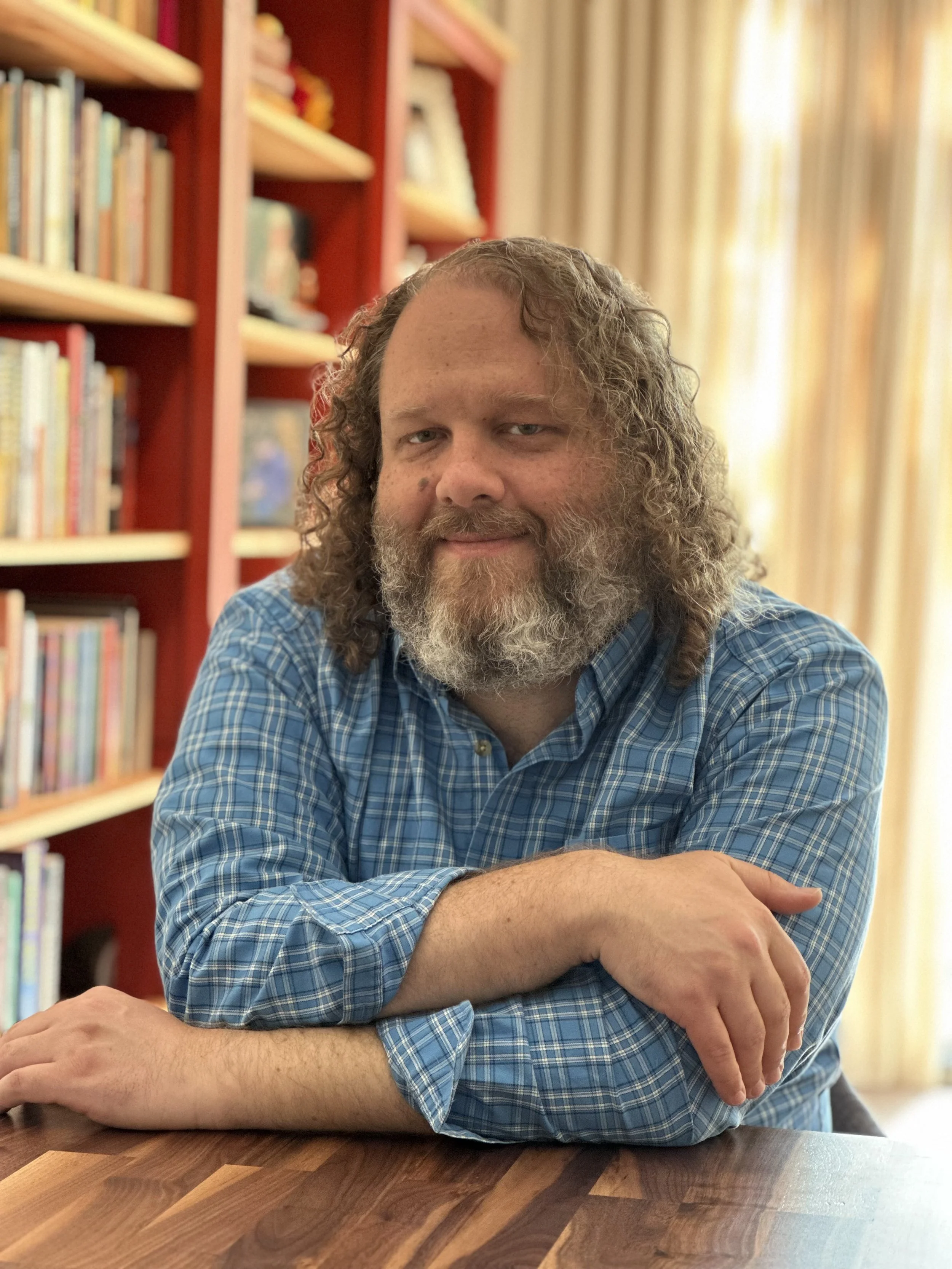 A man with long curly hair and a beard sitting at a wooden table, smiling with his arms crossed. Behind him are bookshelves filled with books and light-colored curtains.