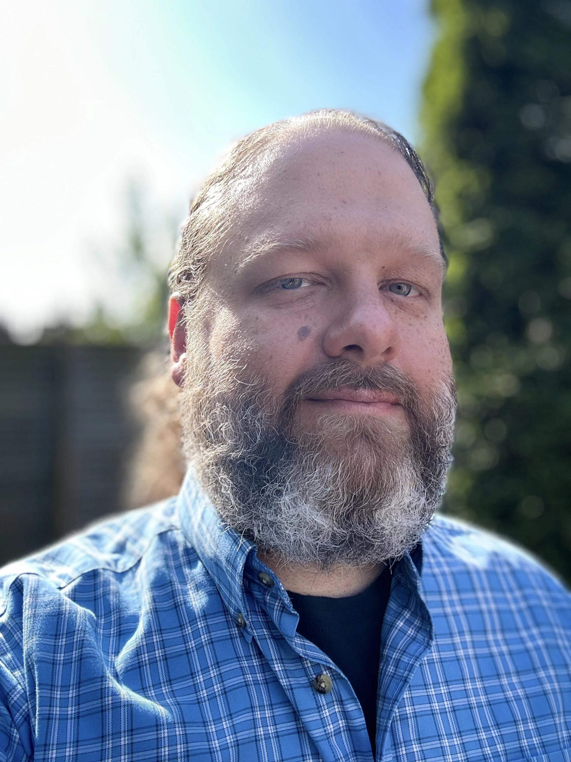 A close-up portrait of a man with a beard and blue eyes, wearing a blue checkered shirt, outdoors on a sunny day with green trees in the background.