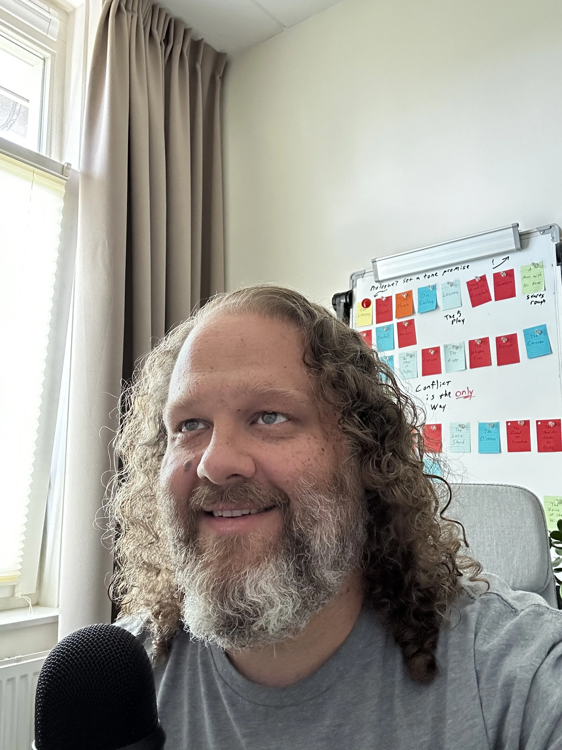 A man with curly hair and a beard smiling while sitting in front of a microphone in a room with white walls, a window with beige curtains, and a whiteboard with colorful sticky notes.