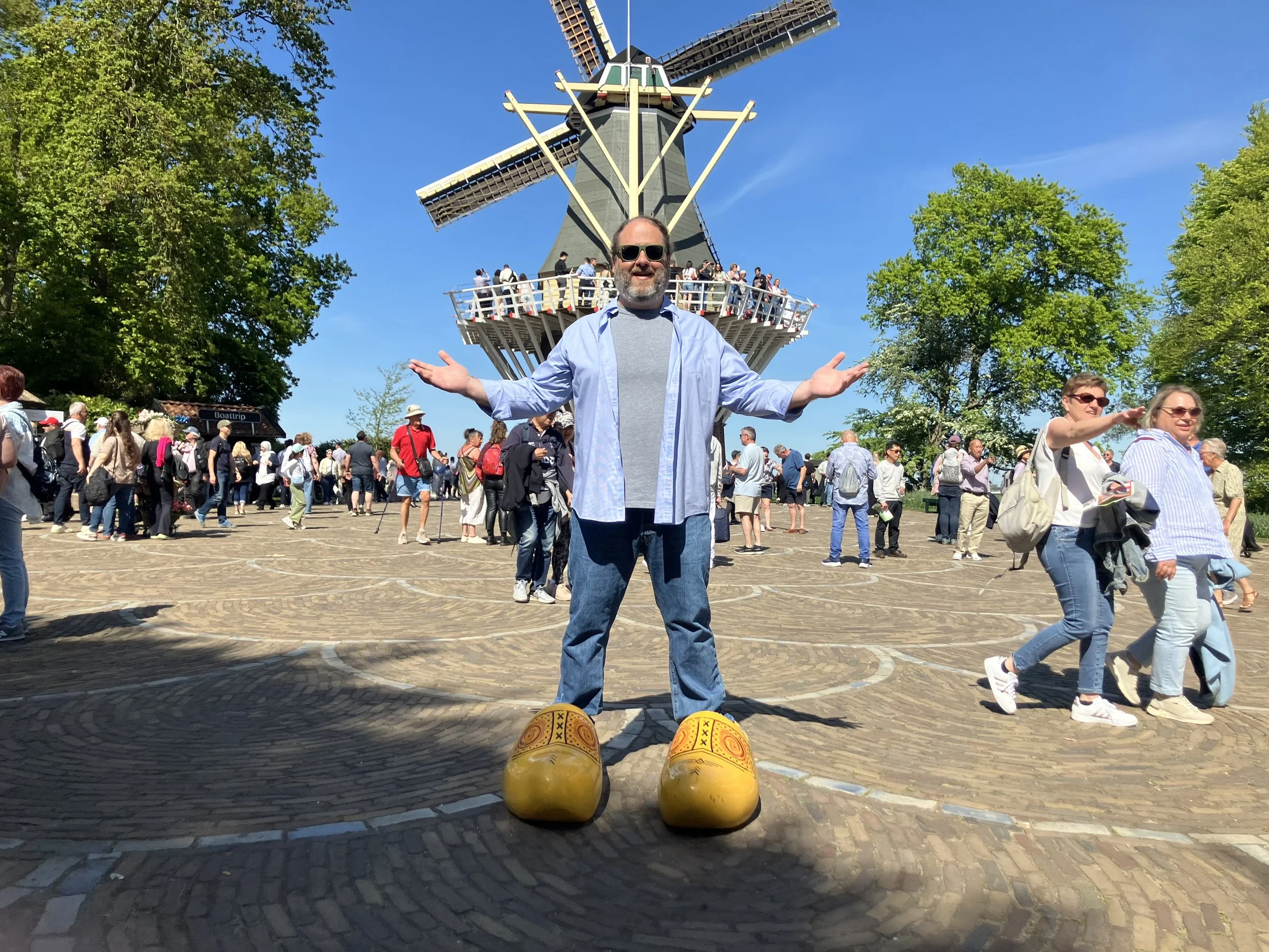 A man wearing sunglasses, a gray t-shirt, and a blue shirt with rolled-up sleeves stands with arms outstretched in front of a large windmill at a busy outdoor tourist site on a sunny day. Many people are walking around and taking pictures.