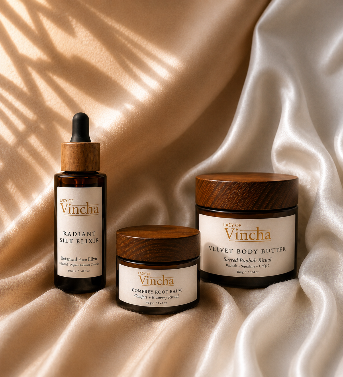 Three skincare products from Lady of Vincha on satin fabric: Radiant Silk Elixir, Comfrey Root Balm, Velvet Body Butter, with wooden caps and white labels.