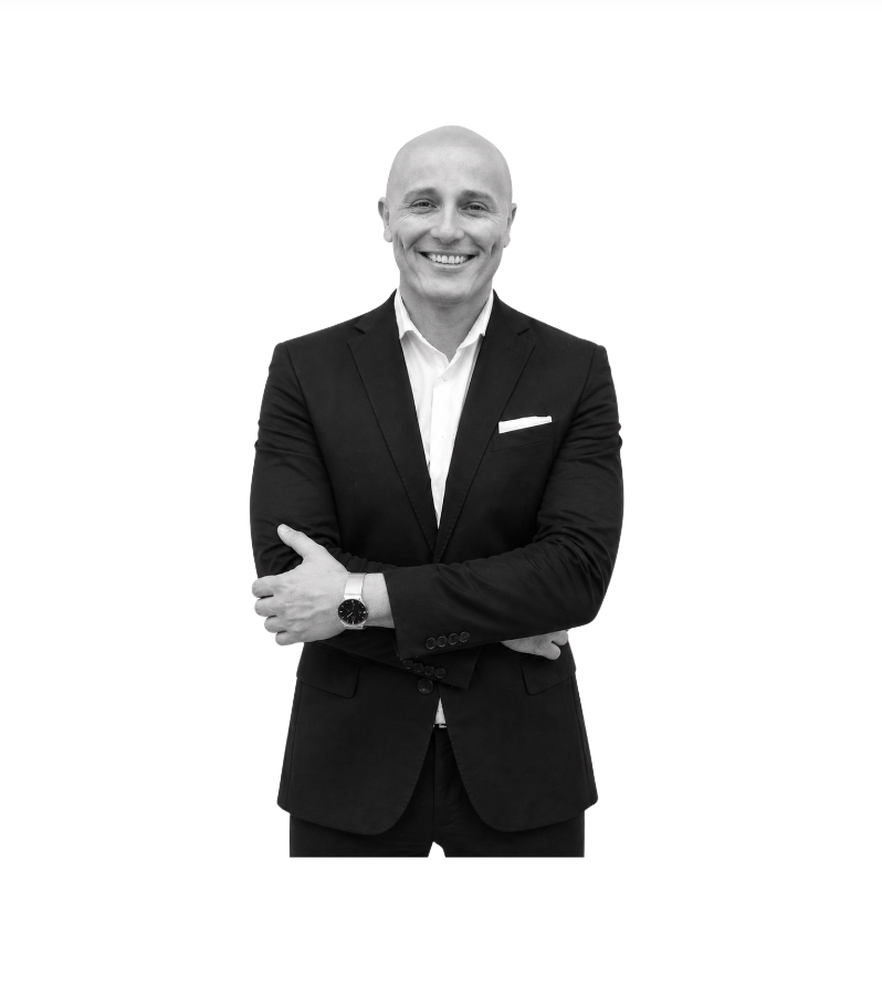 Black and white photo of a smiling man in a black suit, white shirt, with his arms crossed, standing against a plain background.