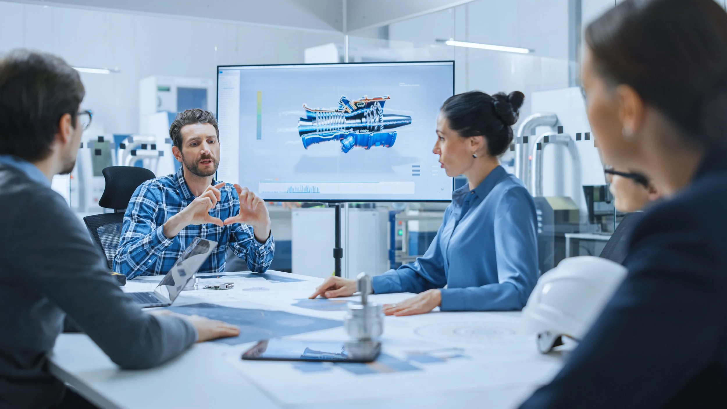 Group of engineers in a meeting discussing a technical diagram of a mechanical part displayed on a large screen in a laboratory or engineering workspace.