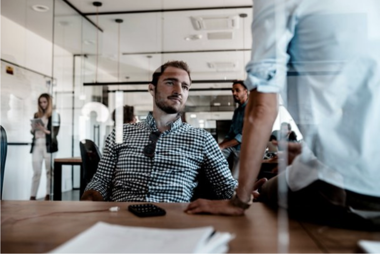 Young man sitting at a conference table, engaged in conversation with a standing person, with others in the background in a modern office setting.