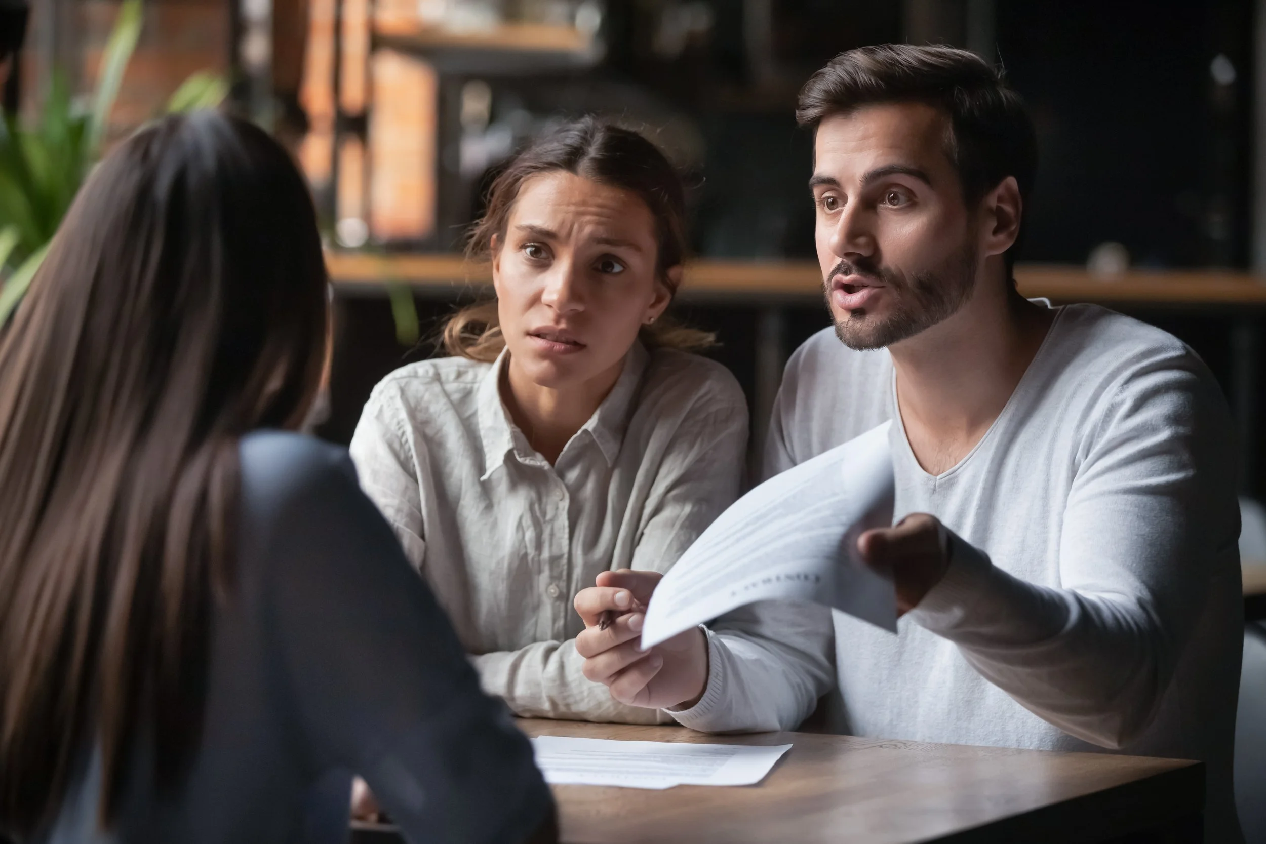 A man and woman having a serious conversation with a young woman in a casual setting, possibly a cafe or office, with documents on the table.