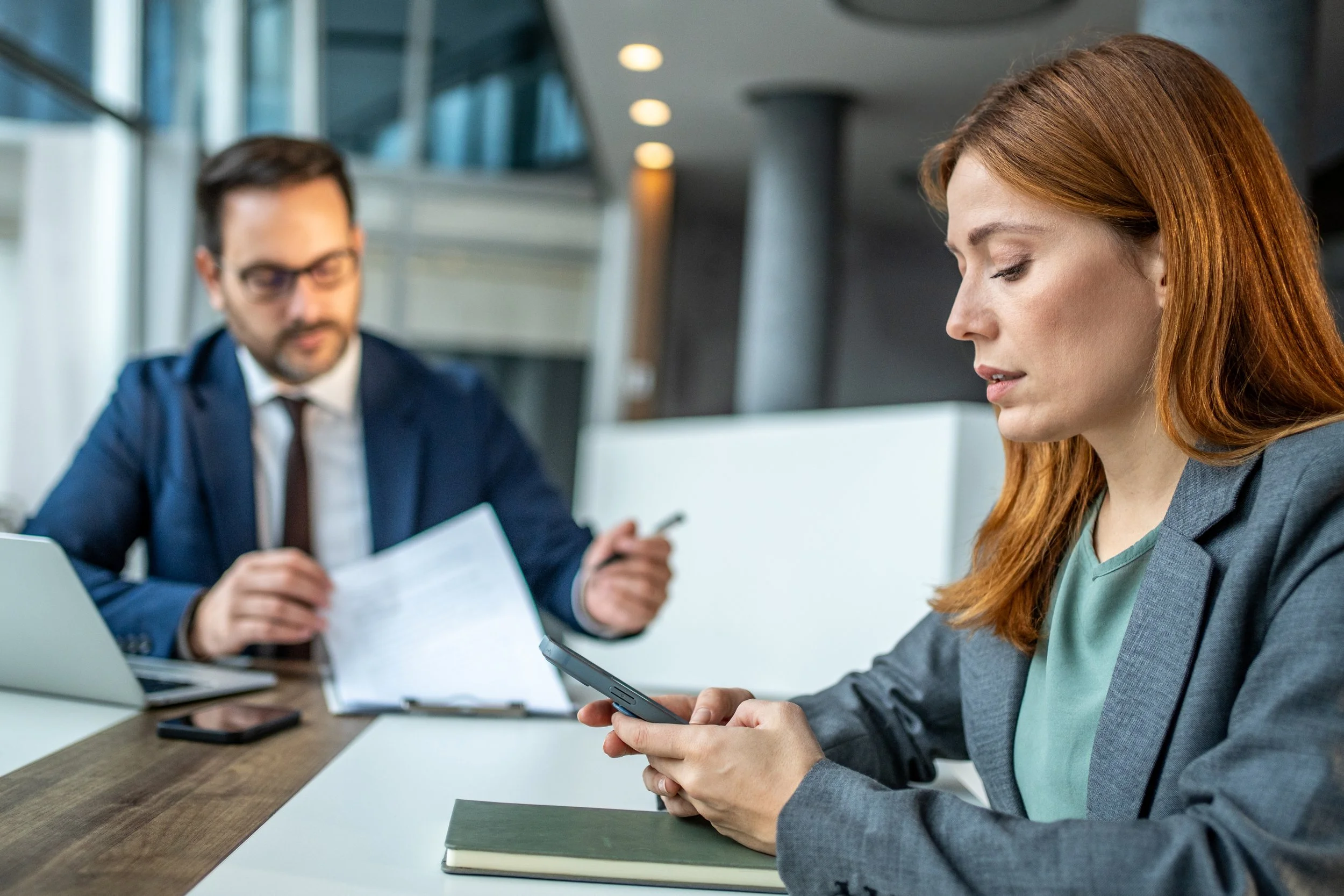 Businesswoman with red hair looking at her phone, seated at a table with a green notebook, in a modern office, with a man in a suit in the background.