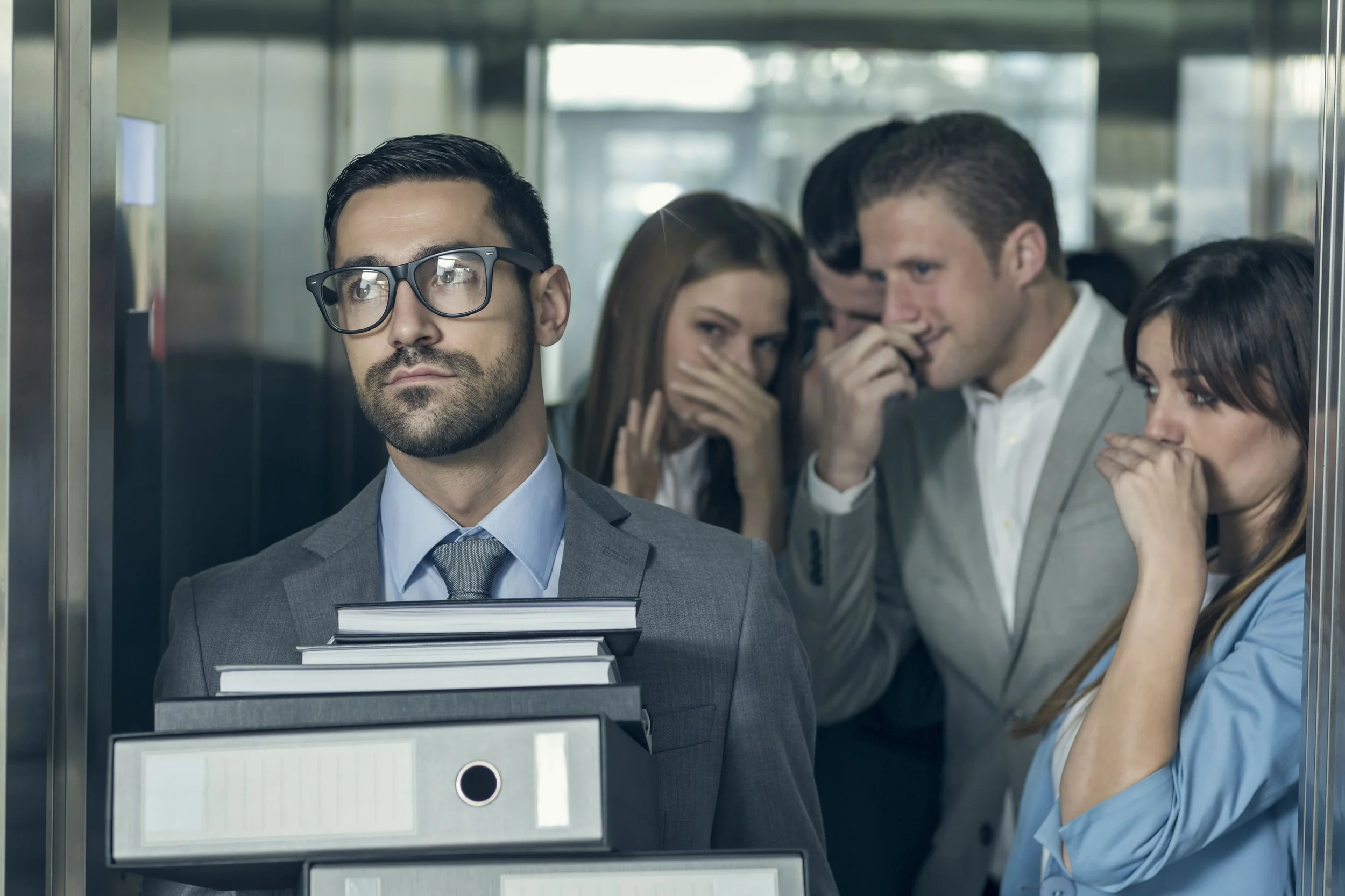 A man in glasses and a suit standing in an elevator, holding several binders and folders, looking serious. Behind him, four other people appear to be whispering or gossiping, with nervous or mischievous expressions.