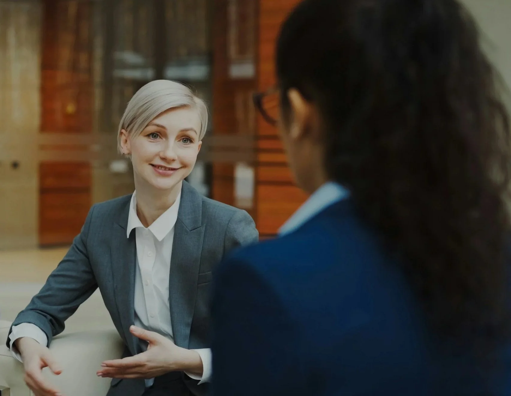 Two women having a conversation in a professional setting. One woman has short gray hair and is wearing a gray blazer, smiling and gesturing with her hands. The other woman has dark hair, glasses, and a blue blazer, facing her.