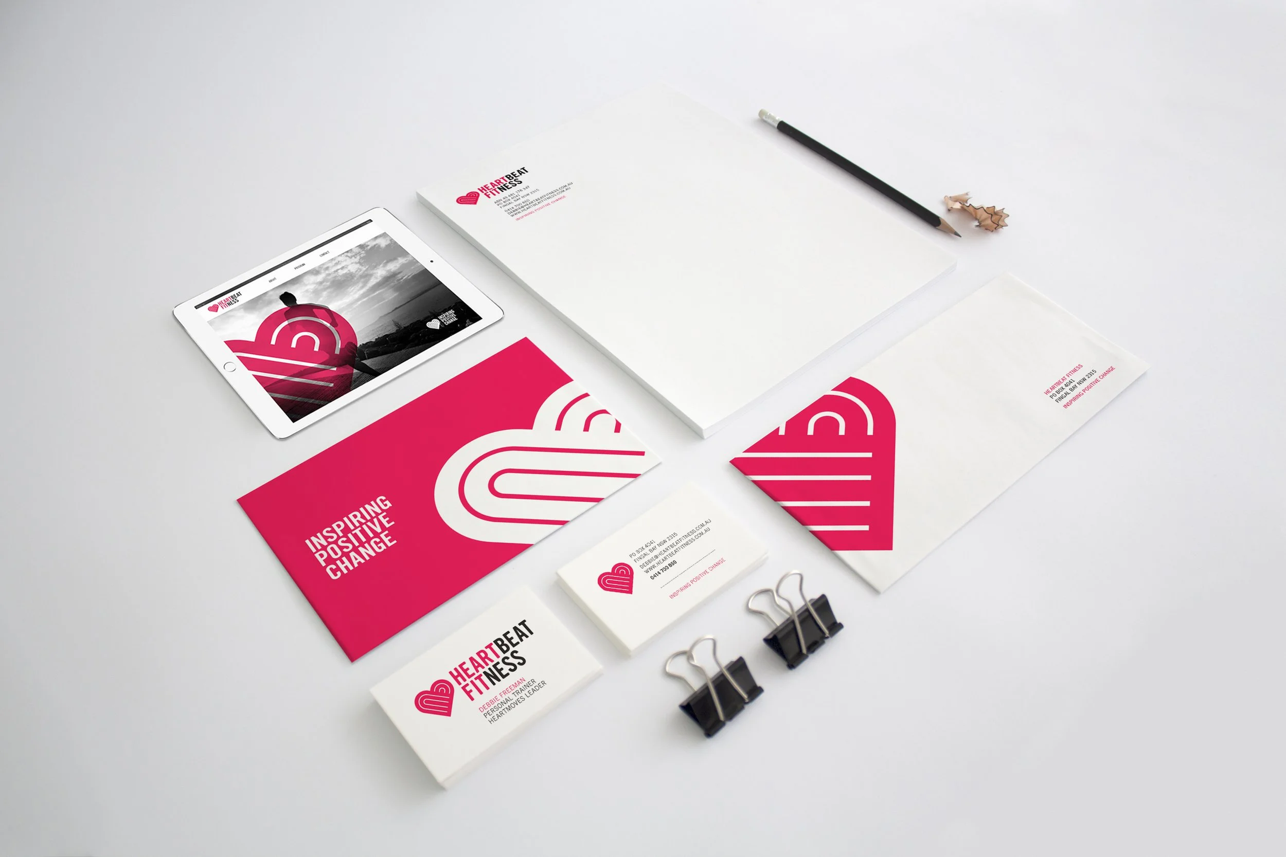 Collection of branding and promotional materials for Heartbeat Fitness, including a tablet displaying a website, flyers and business cards with the Heartbeat Fitness logo and the slogan 'Inspiring Positive Change', a pencil, paper clips, and a small pile of pencil shavings on a white surface.