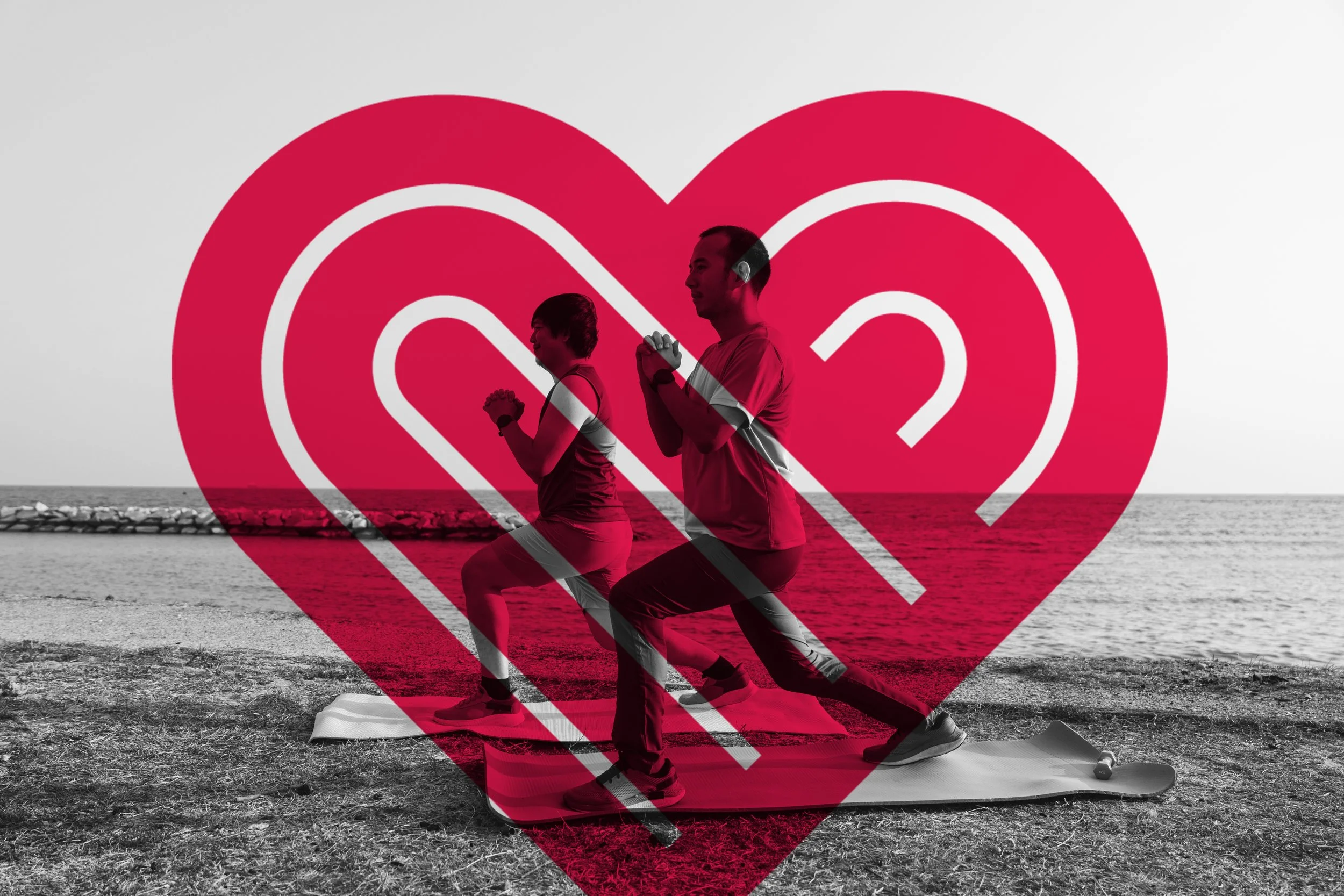 Two people practicing yoga on a beach, kneeling on yoga mats with hands in prayer position, large red heart shape overlaying the image.