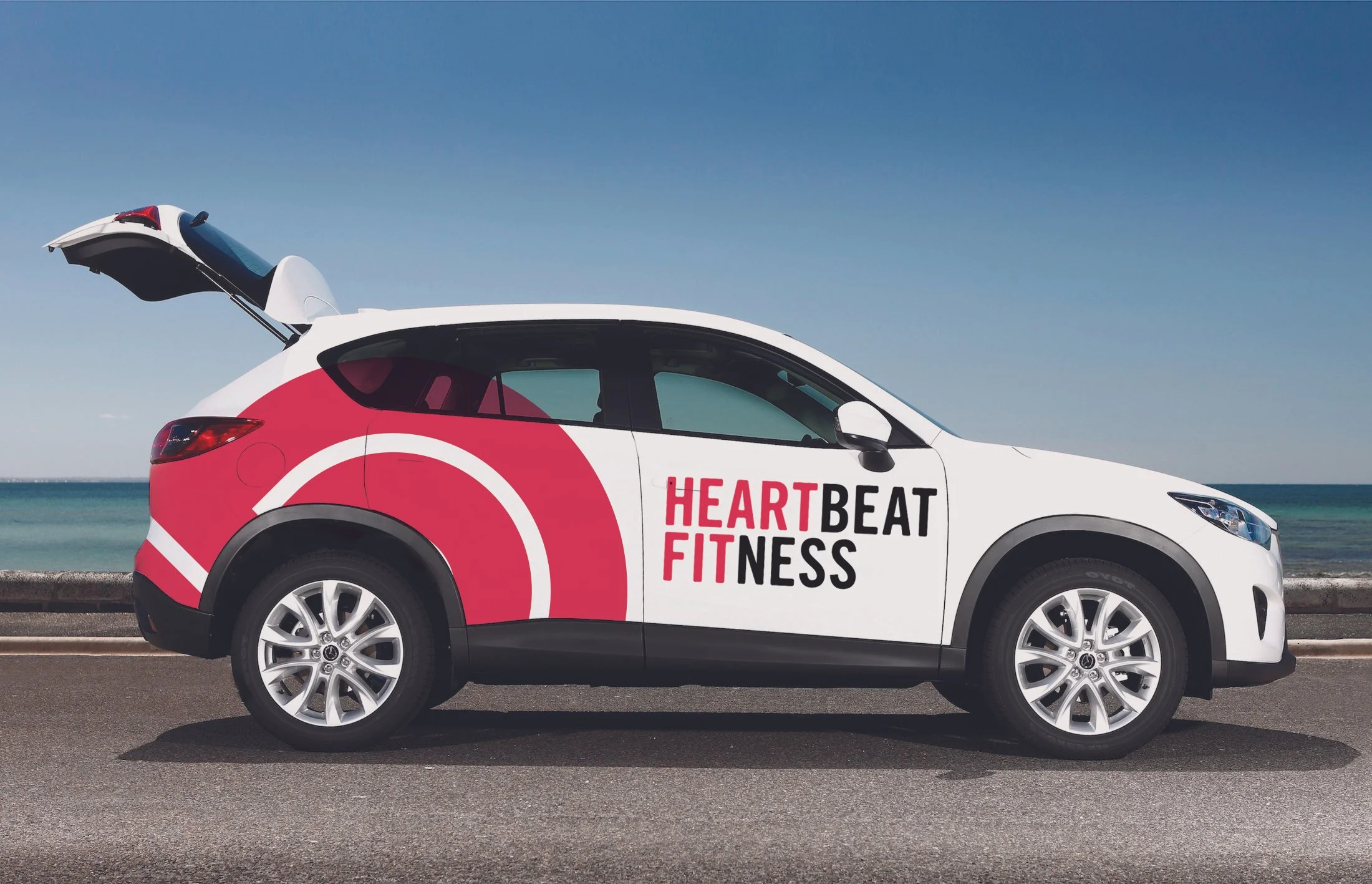 A white car with red and black graphics and the words 'Heartbeat Fitness' on the side, parked by a beach with a blue sky and ocean in the background.