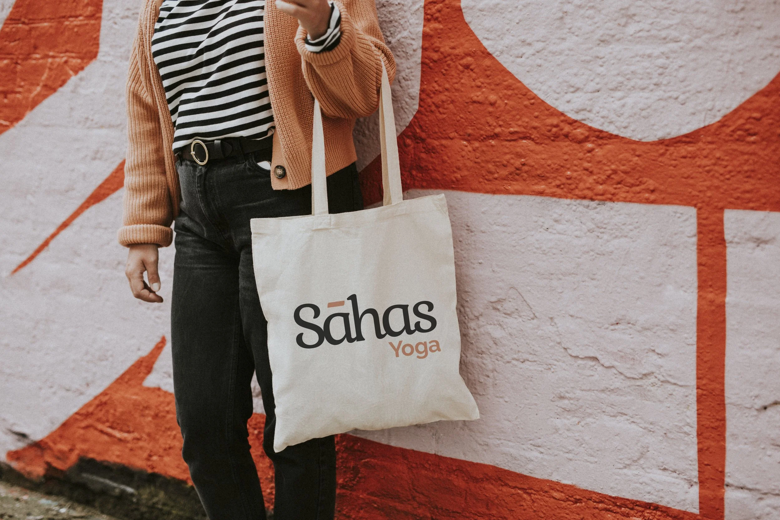 Person holding a tote bag with the text 'sahas Yoga' standing against a colorful wall with red, white, and orange mural patterns.