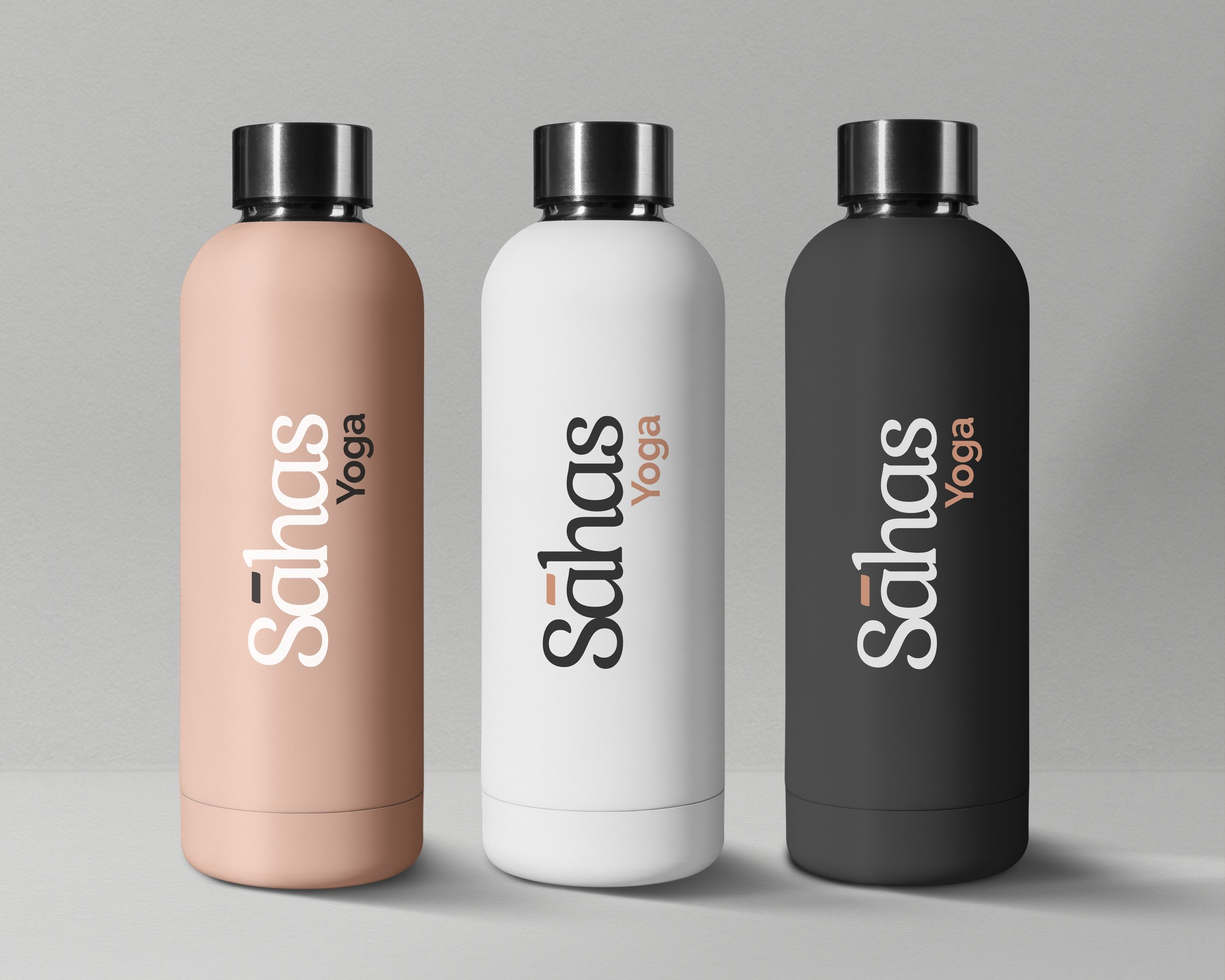 Three stainless steel water bottles with black caps, in pink, white, and black, placed on a gray surface against a gray wall, with the brand name 'Acnus Yoga' printed vertically.