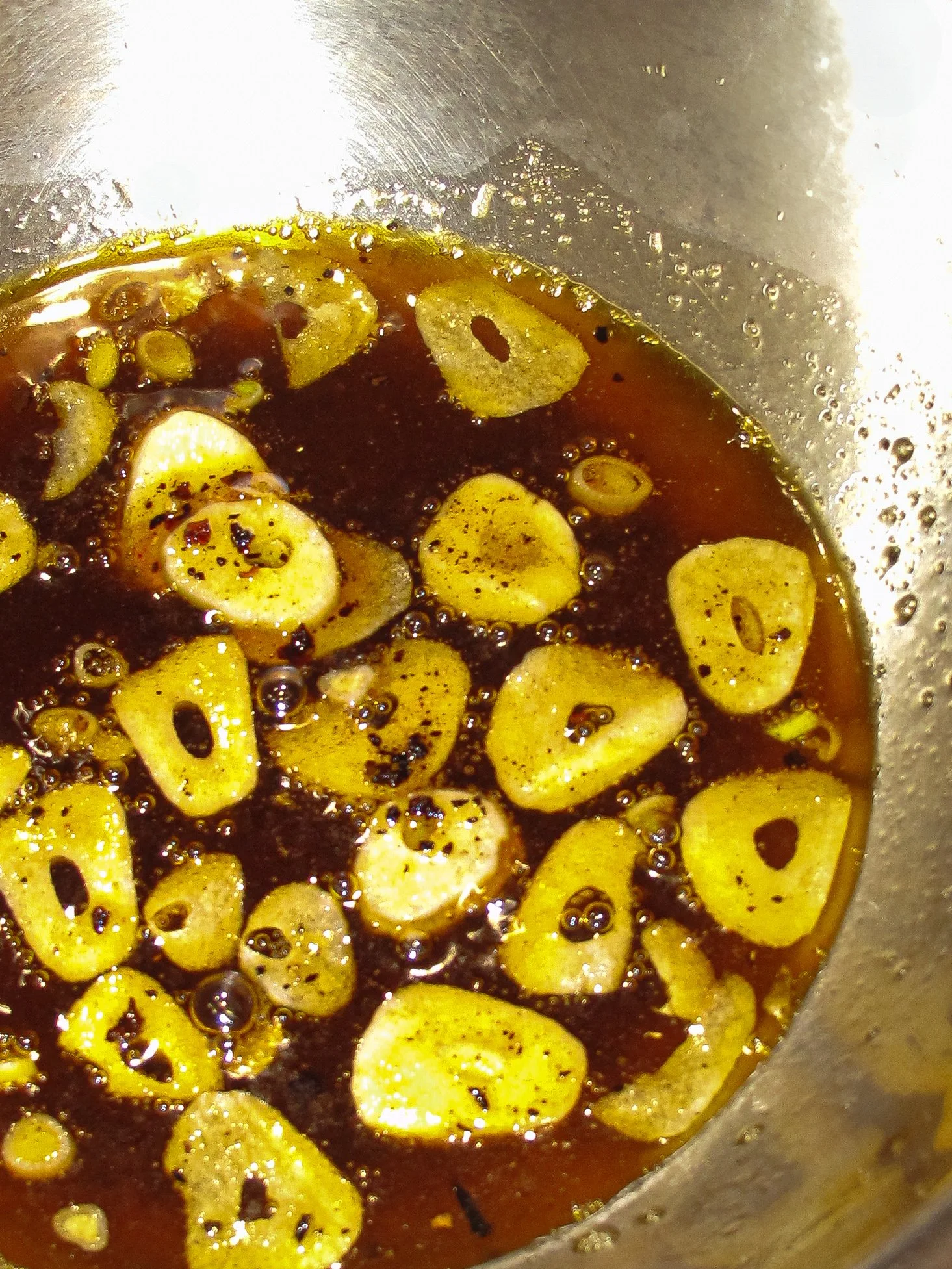 Sliced garlic cloves cooking in a dark sauce or oil in a metallic pan.