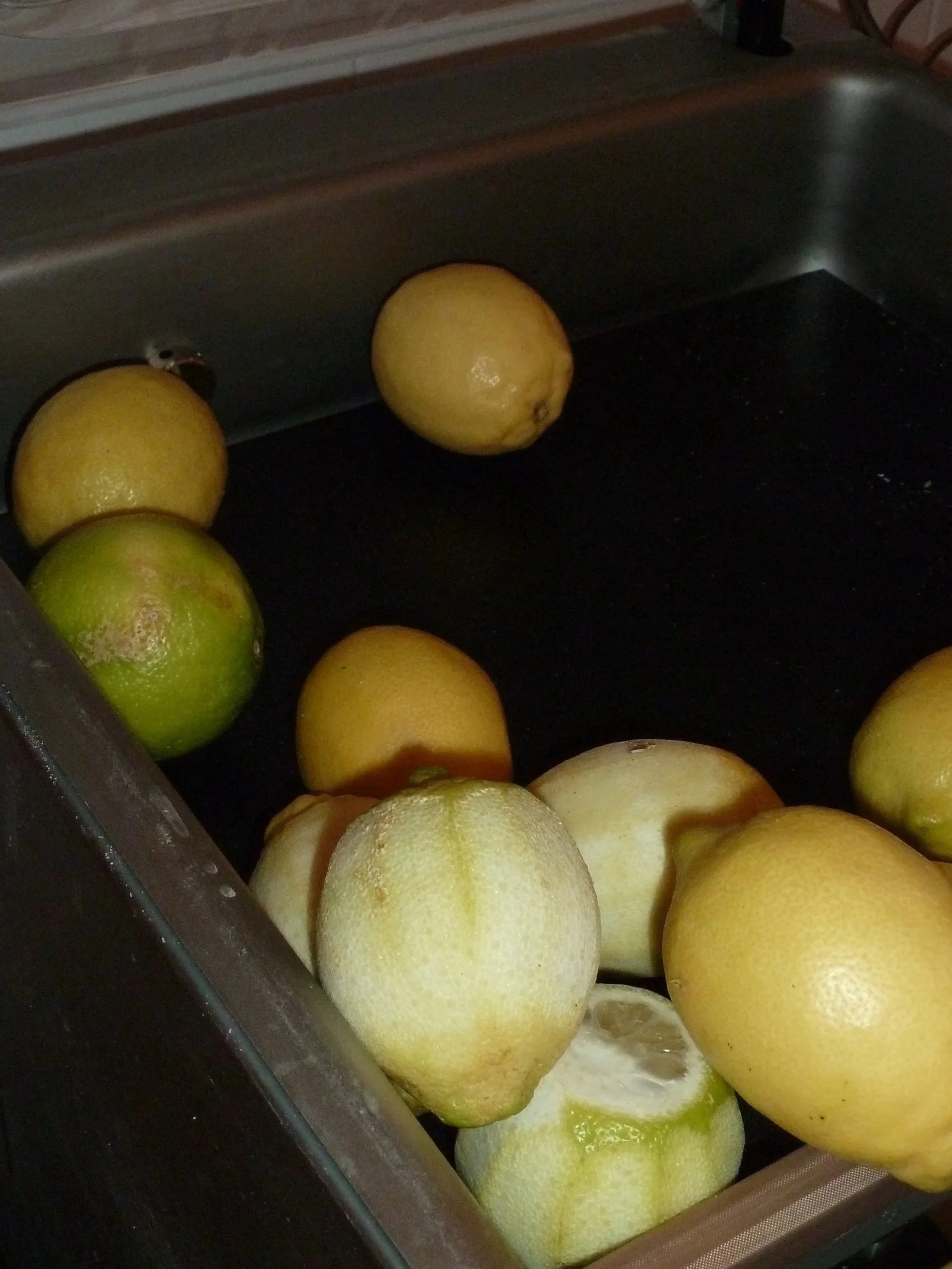 A bunch of lemons in a tray, some are yellow and some are green, with a black surface underneath.