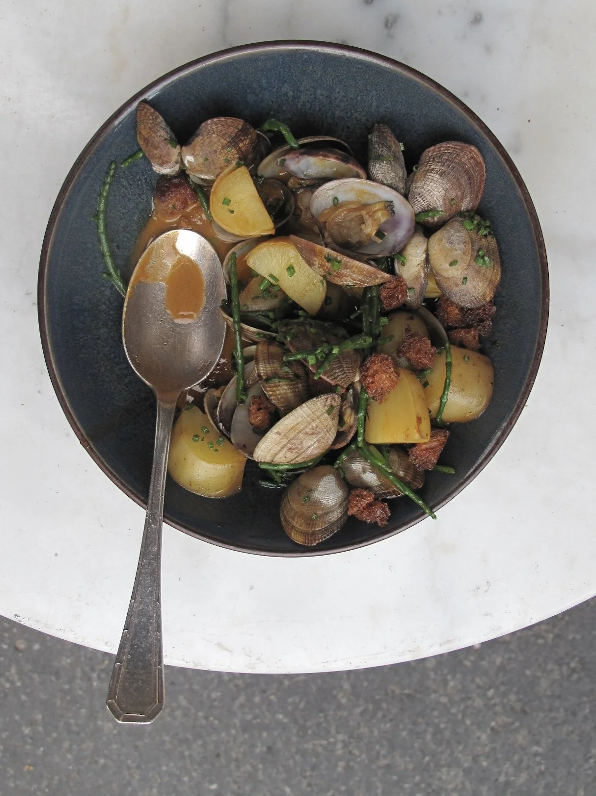 A bowl of cooked clams with potatoes, green beans, and chopped herbs, with a spoon resting inside on a white surface.