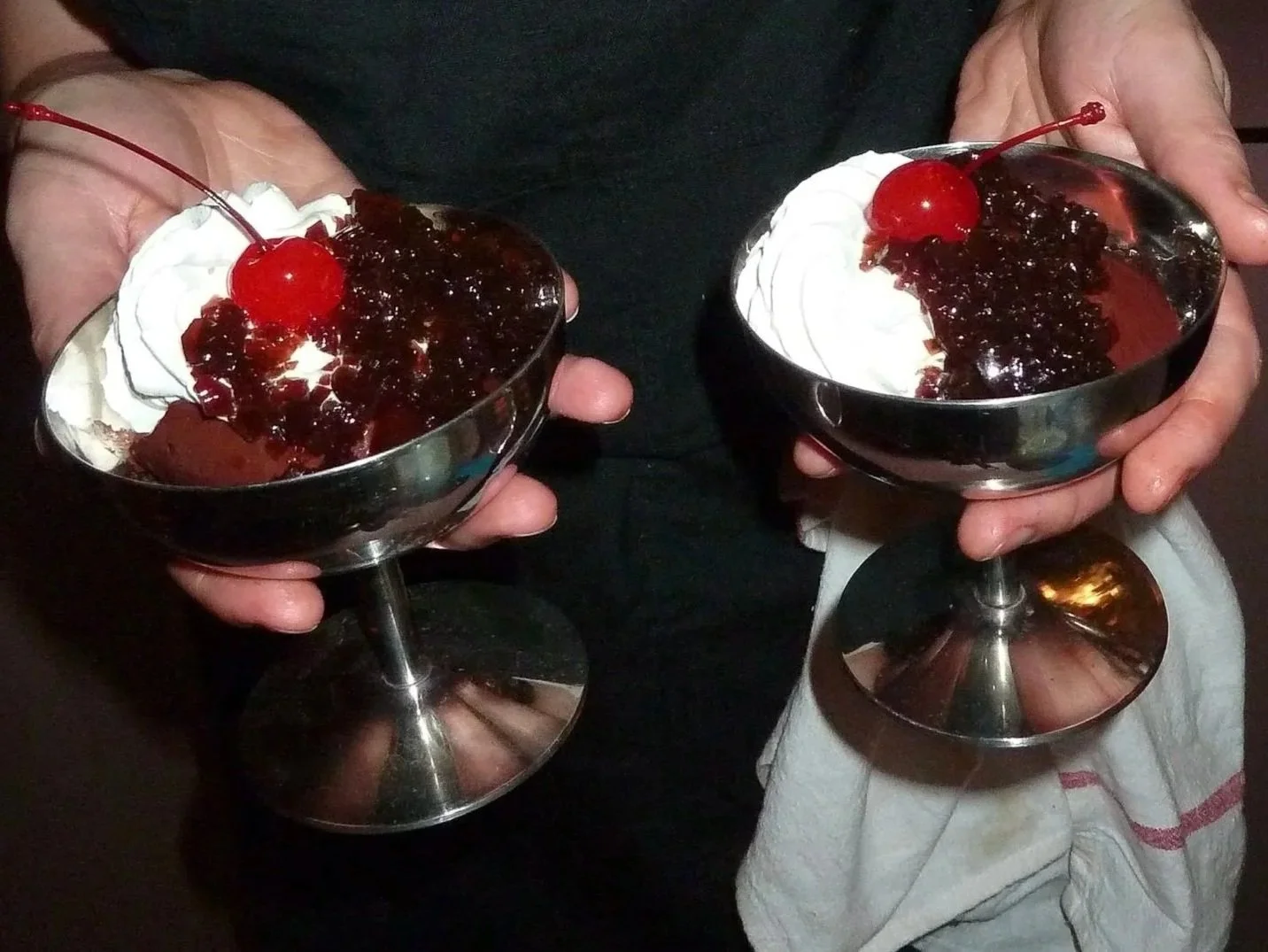 Two people holding metal bowls of dessert topped with cherries, whipped cream, and cherry sauce.