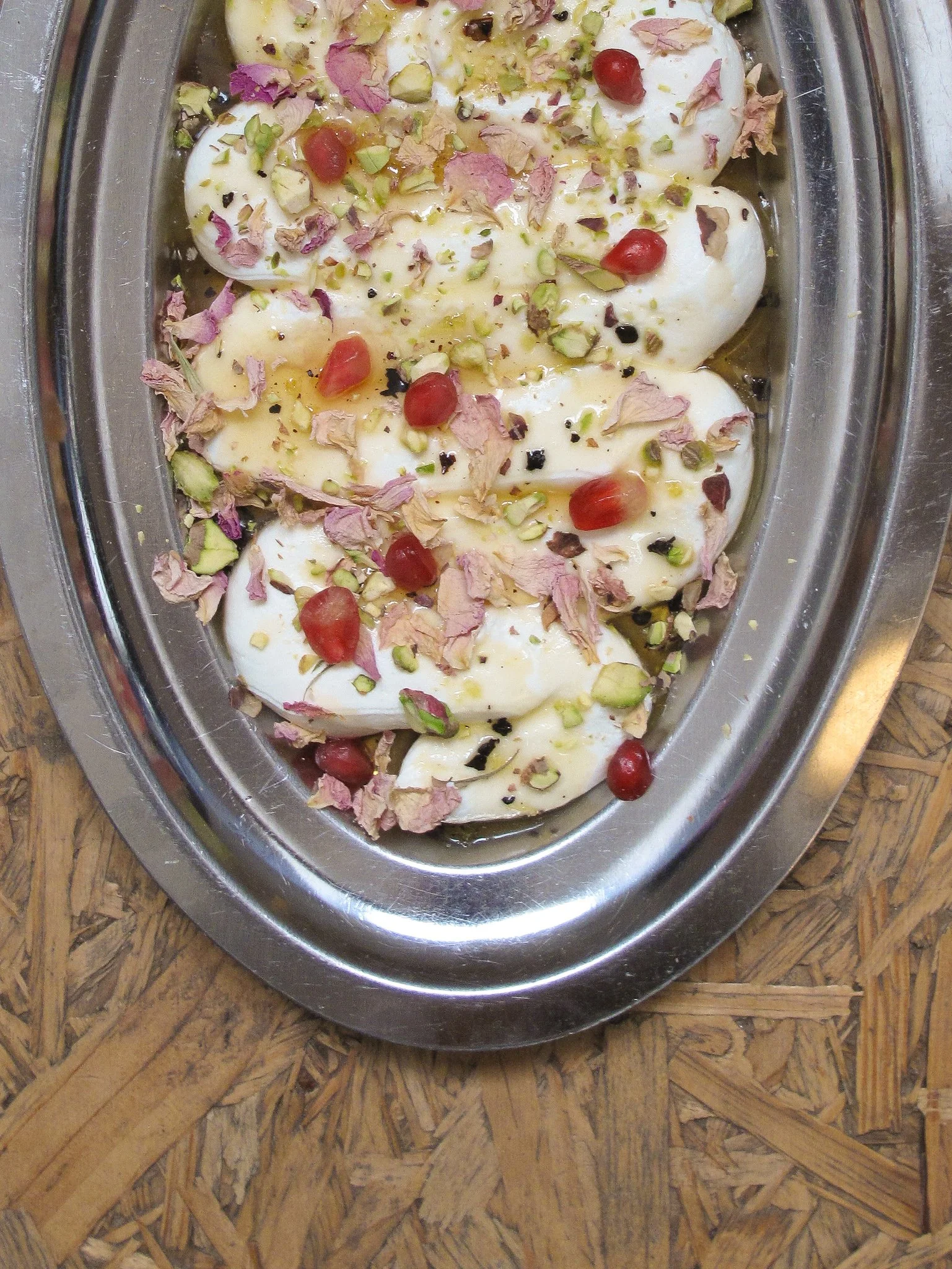 Labneh garnished with chopped pistachios, pomegranate seeds, dried rose petals, and ground black pepper, served on a metal platter.