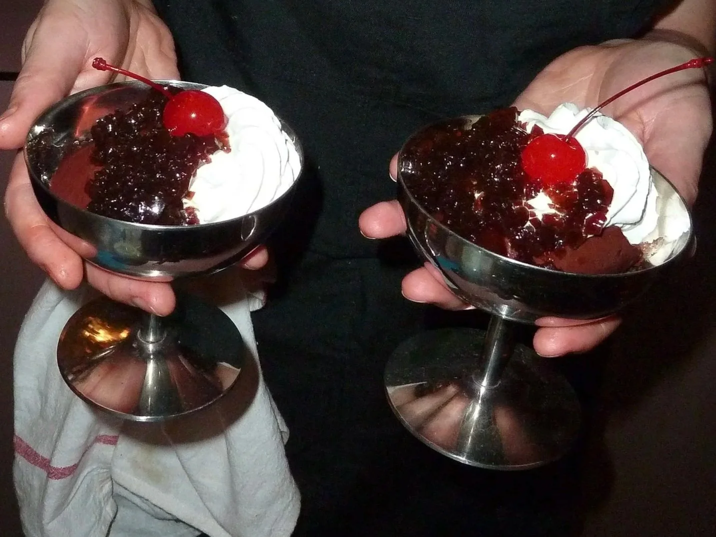 Two people holding metal dessert bowls filled with ice cream topped with cherry and berry compote and whipped cream.