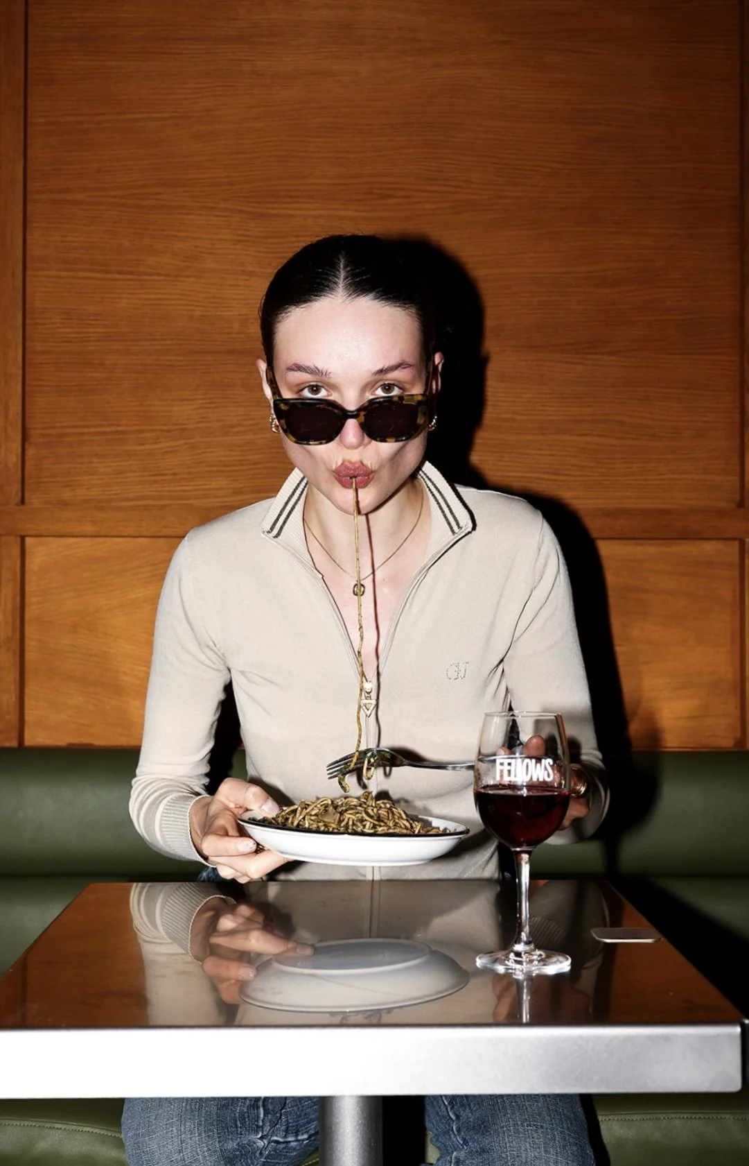 Emma KONC, is eating a plate of spaghetti and drinking red wine at a restaurant. She has a playful expression with her lips pursed and a piece of spaghetti hanging from her mouth.