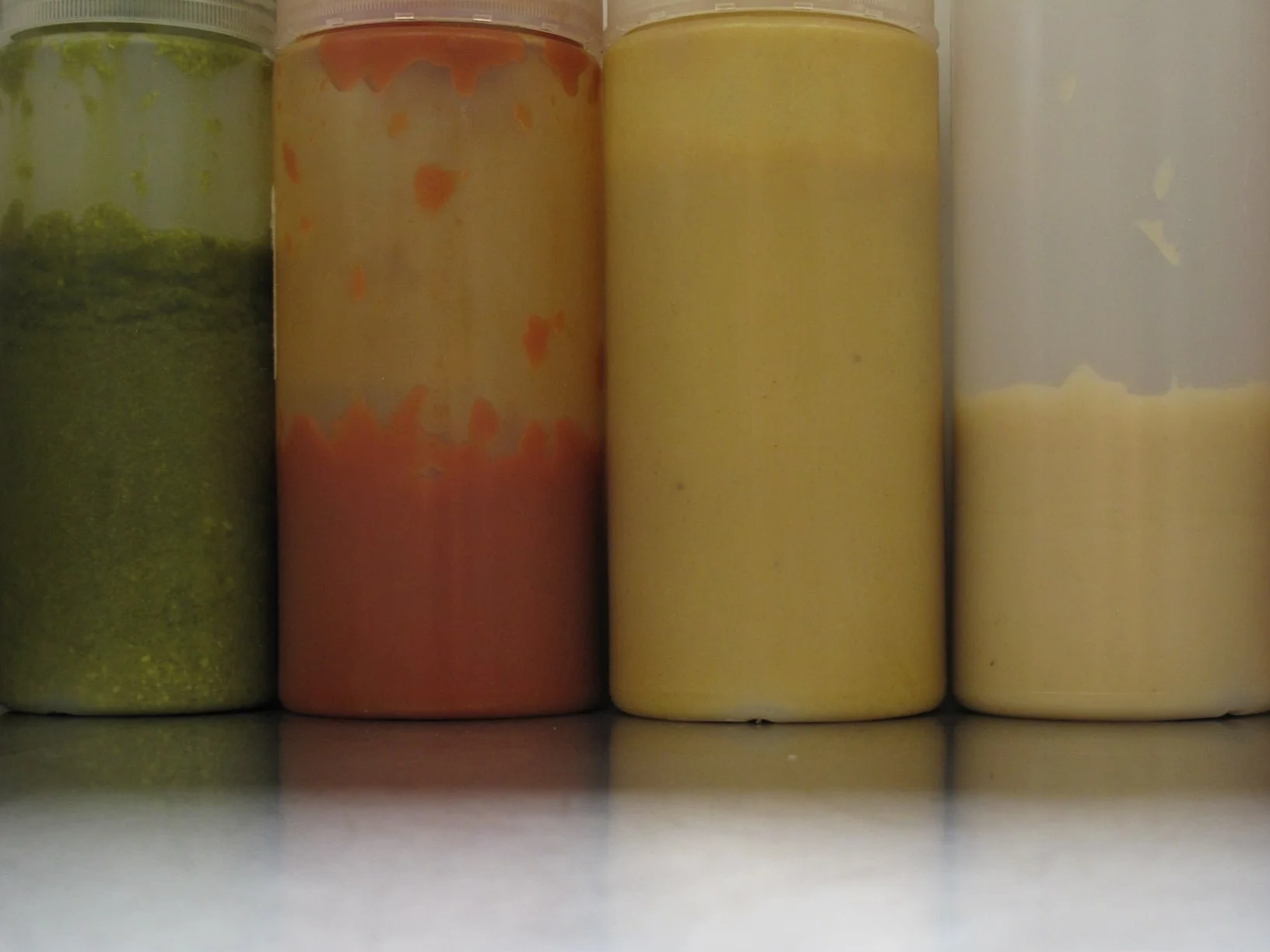 Four bottles of different colored sauces lined up on a shelf, taken from a low angle.