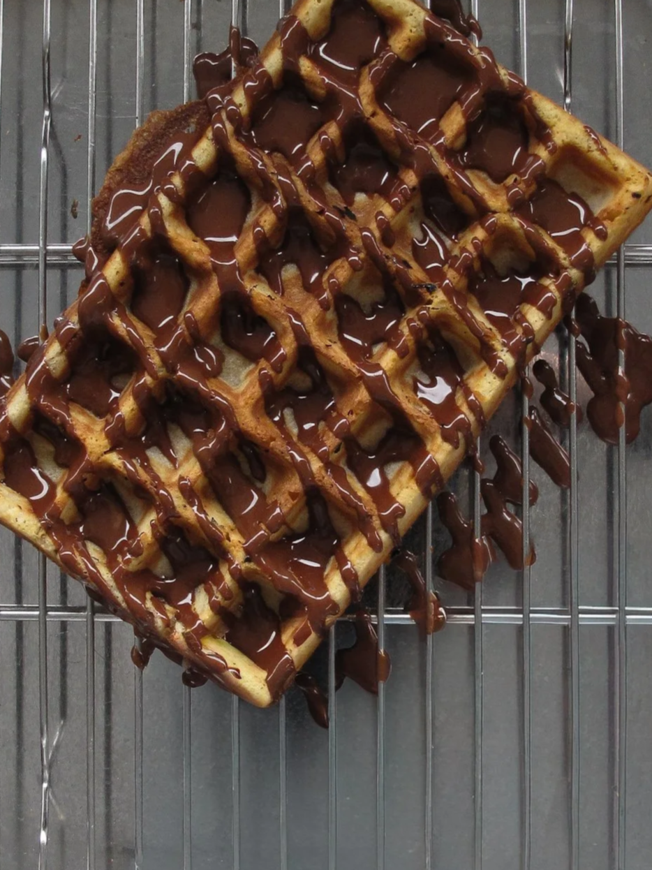 A golden-brown Belgian waffle drizzled with melted chocolate on a wire rack with chocolate spills on the surface underneath.