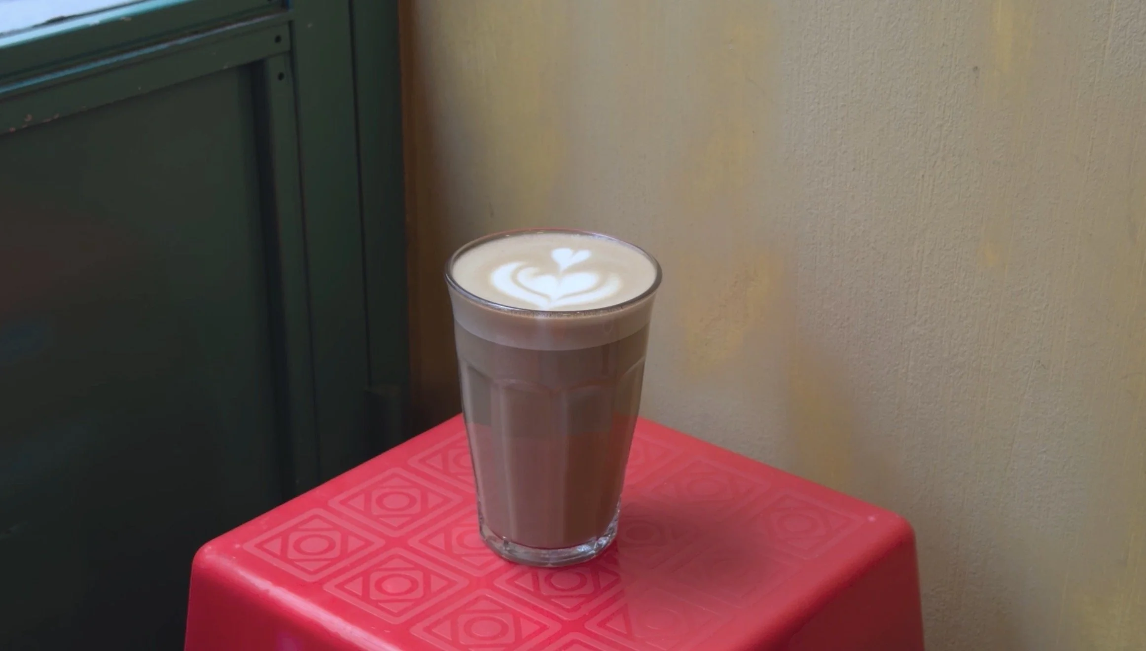 A tall glass of latte with milk foam and latte art on top, placed on a red stool against a beige wall and green cabinet.