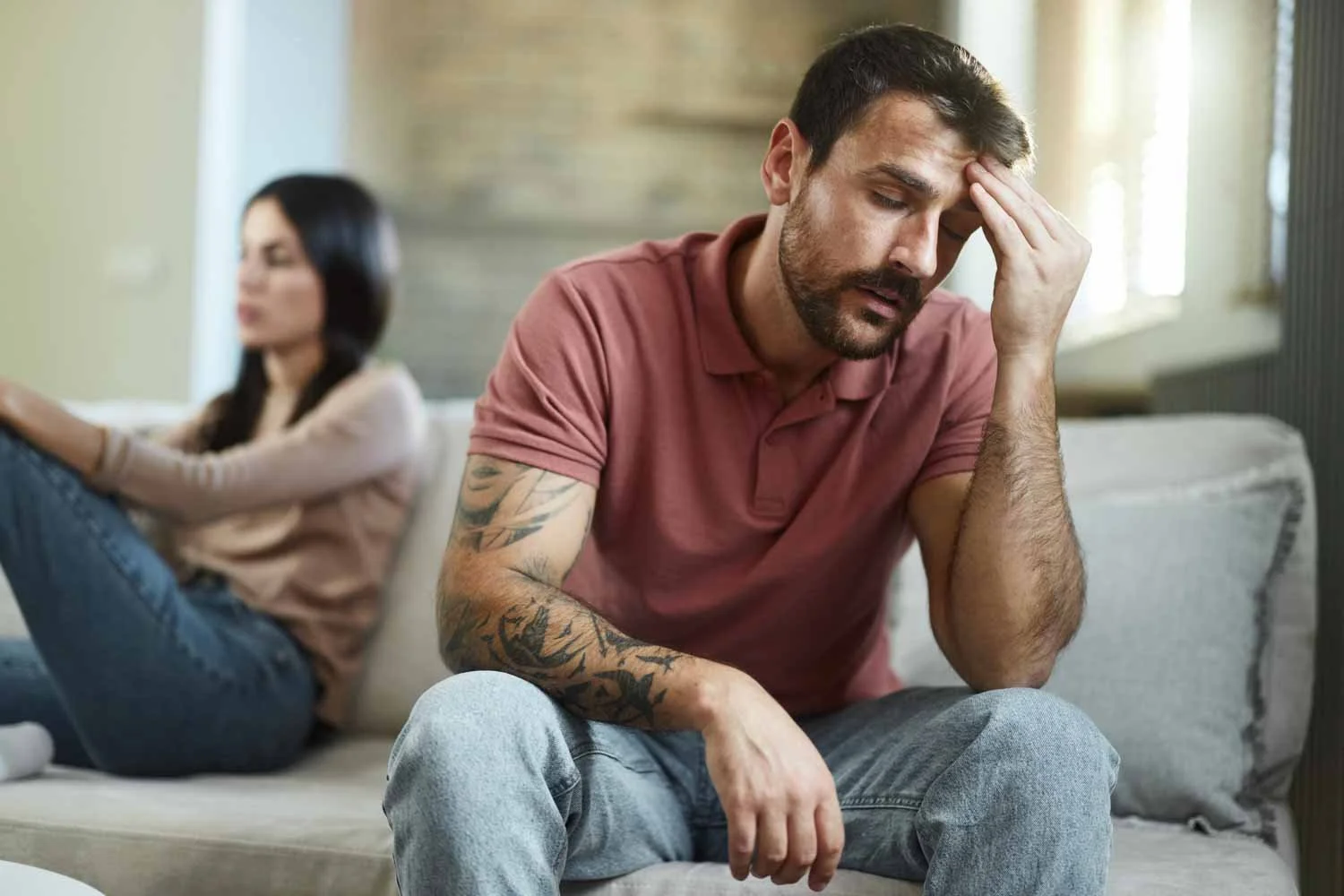 En man ser ut att vara stressed eller bekymrad, sitter på en soffa med handen på huvudet, medan en kvinna i bakgrunden verkar oberörd. Paul Howard, psykolog.