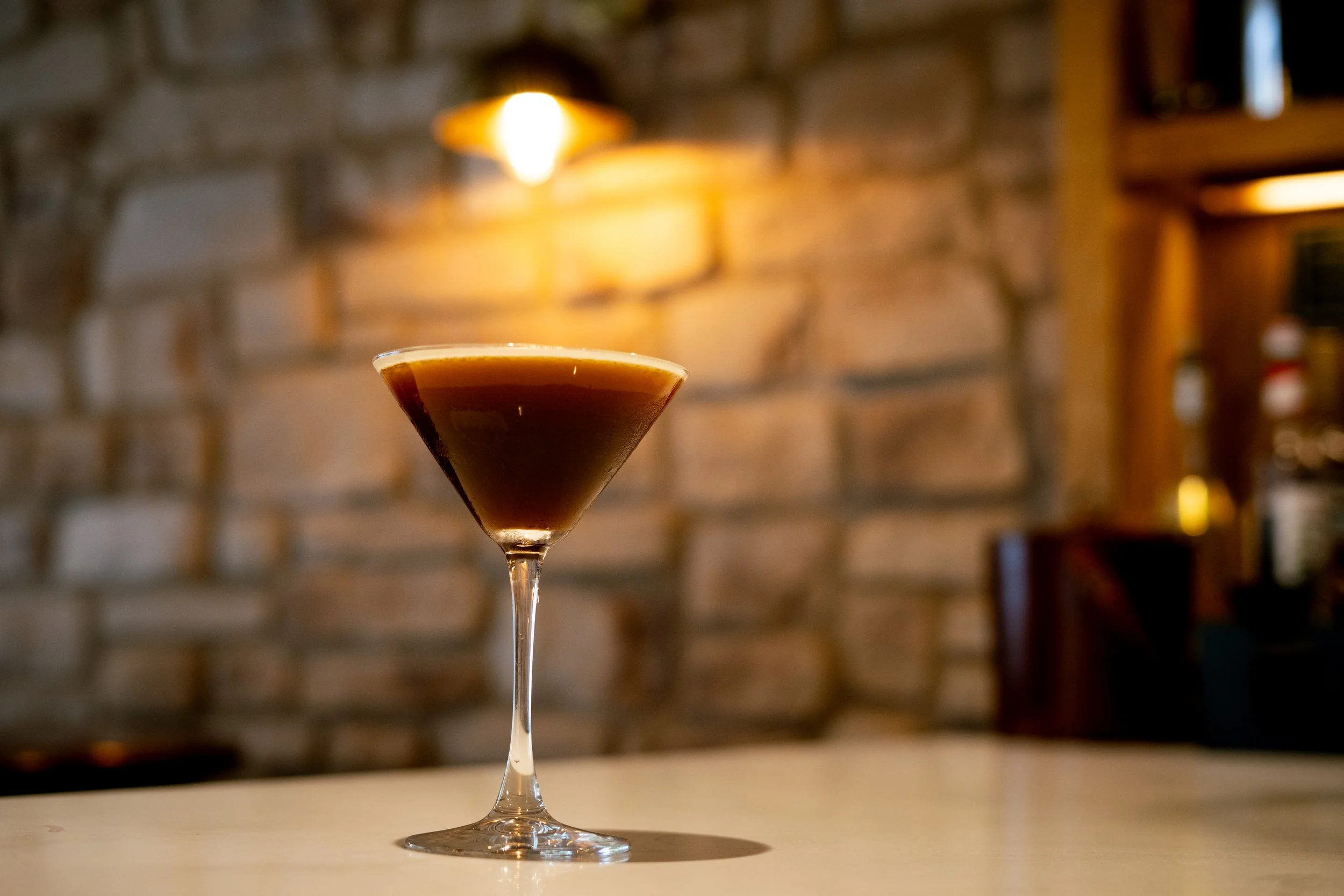 A cocktail glass filled with a dark brown drink, on a white surface with a brick wall and warm lighting in the background.