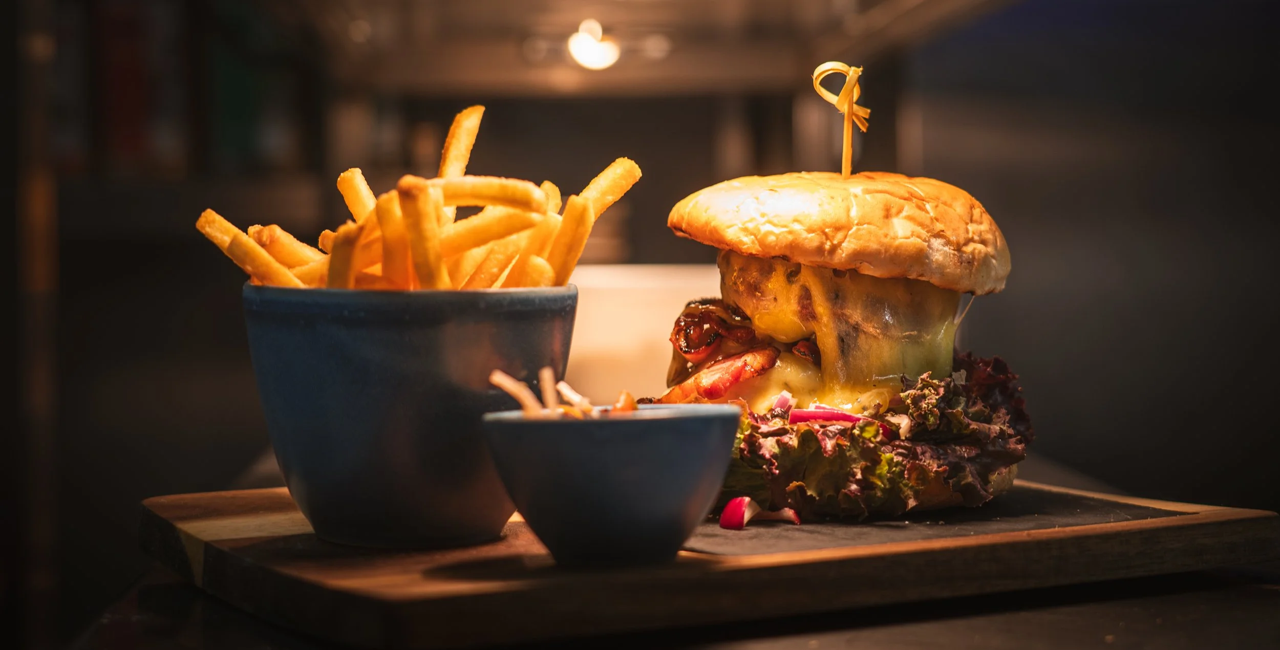 A cheeseburger with melted cheese, lettuce, and bacon, served with a side of French fries in a blue bowl on a wooden serving board.