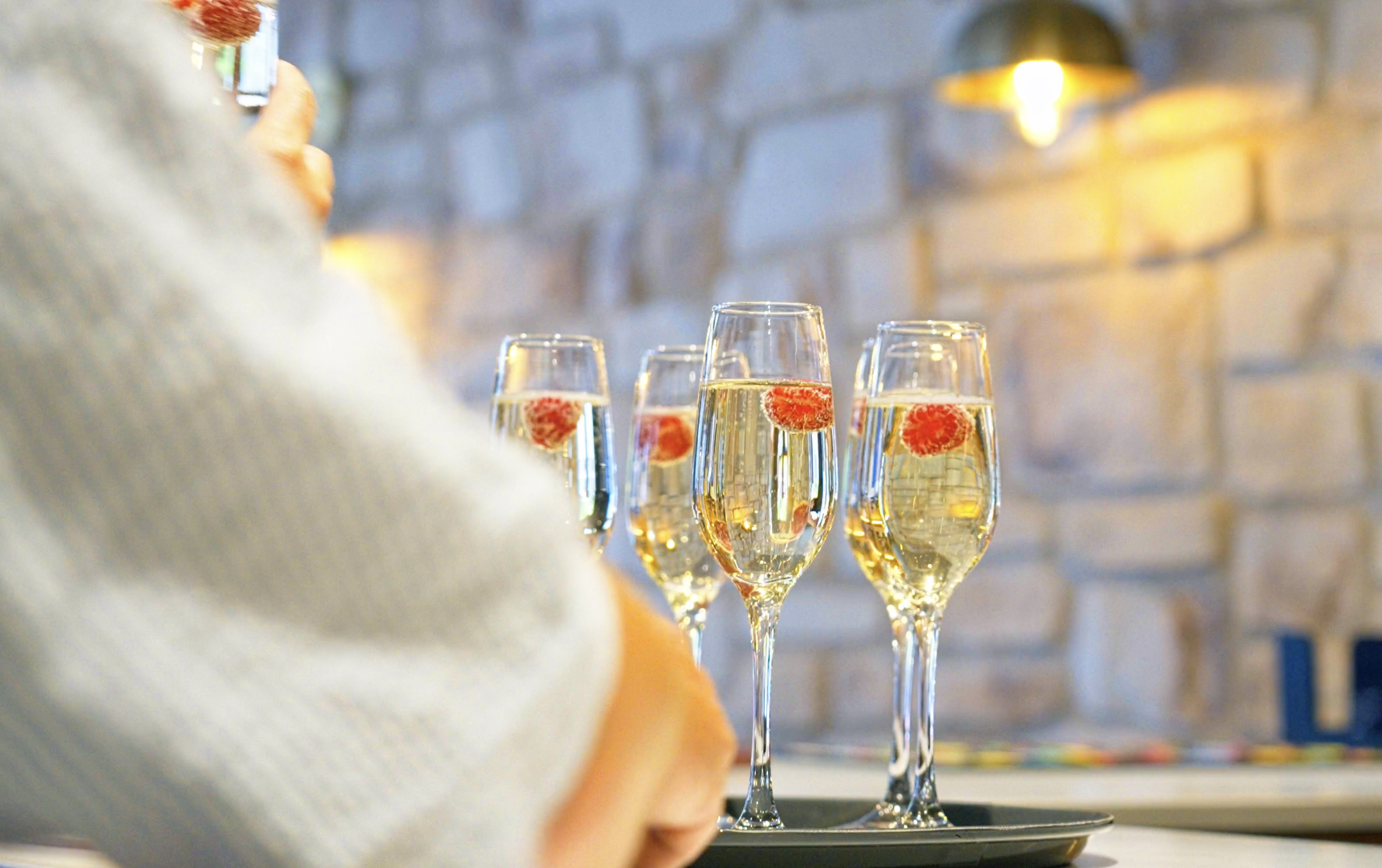 Four glasses of champagne with floating raspberries on a black tray, blurred brick wall and warm lighting in the background, person holding a glass in the foreground.